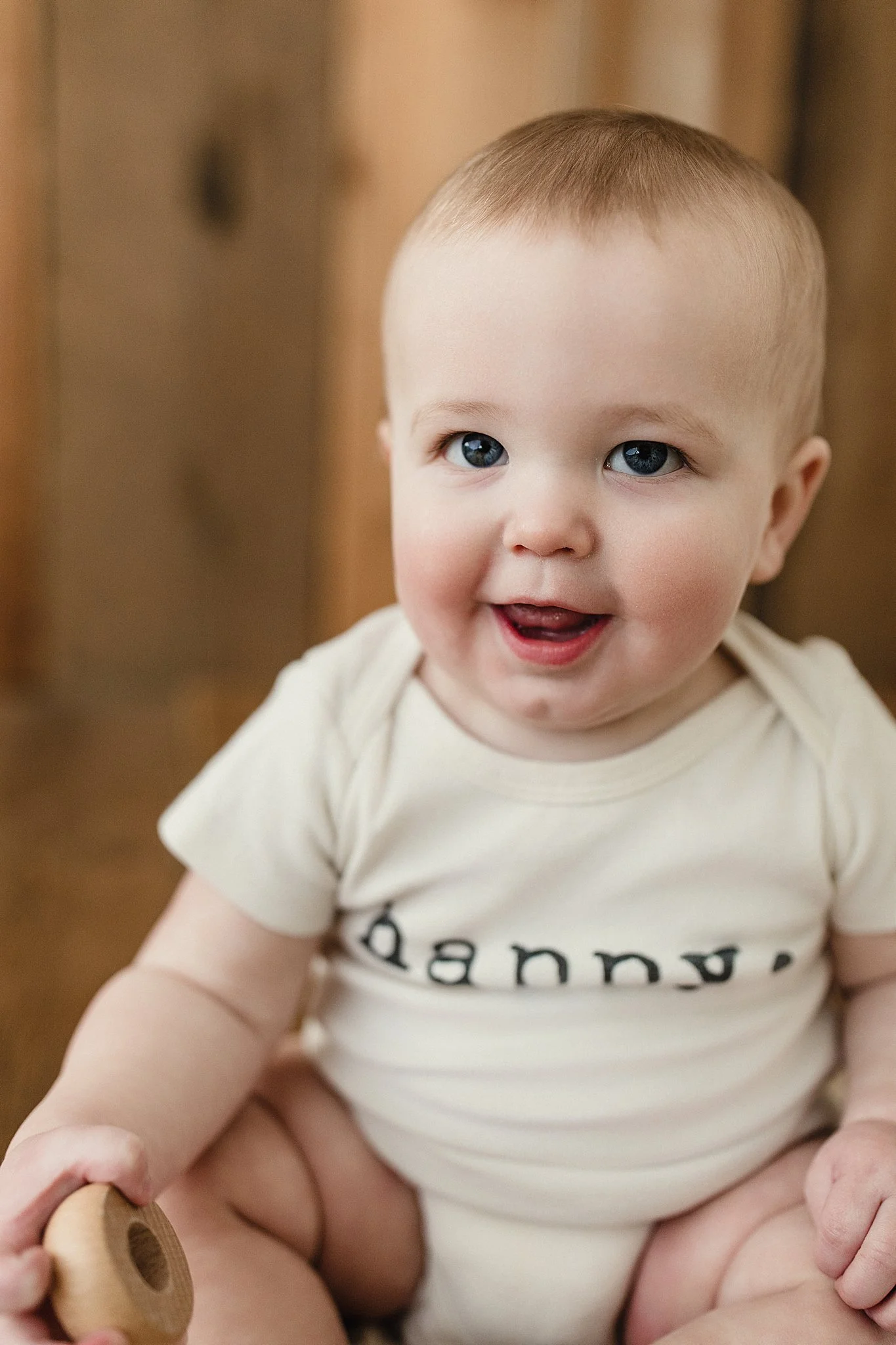 A smiling baby sitting on a wooden surface, wearing a cream-colored shirt and holding a small object during a milestone photoshoot for a first birthday in Naperville.