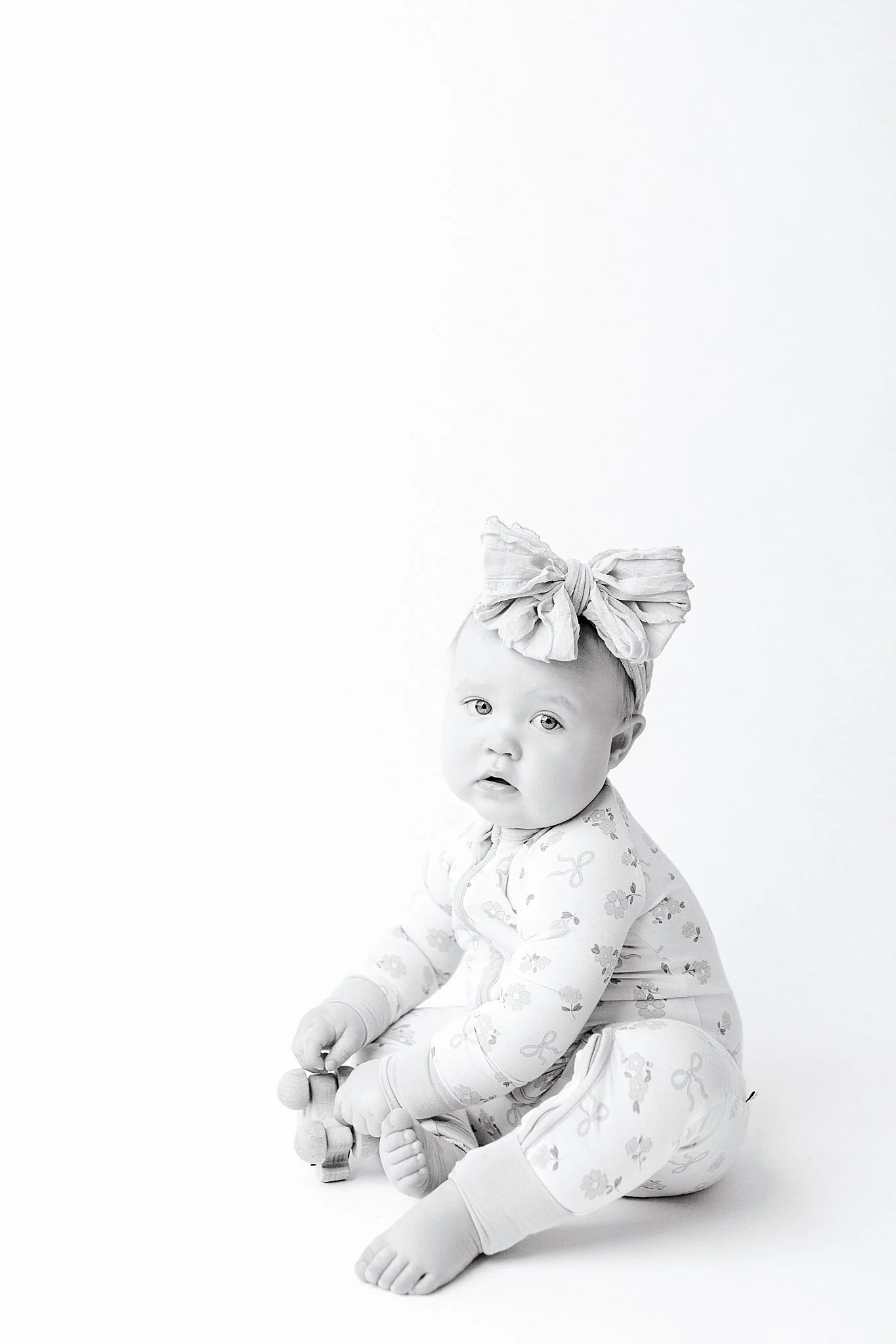 A baby girl with light-colored eyes and a bow headband, sitting on the floor in a onesie with a bow and flower pattern, holding a small toy, in a bright, white, minimal studio setting for baby milestone photos in Naperville.