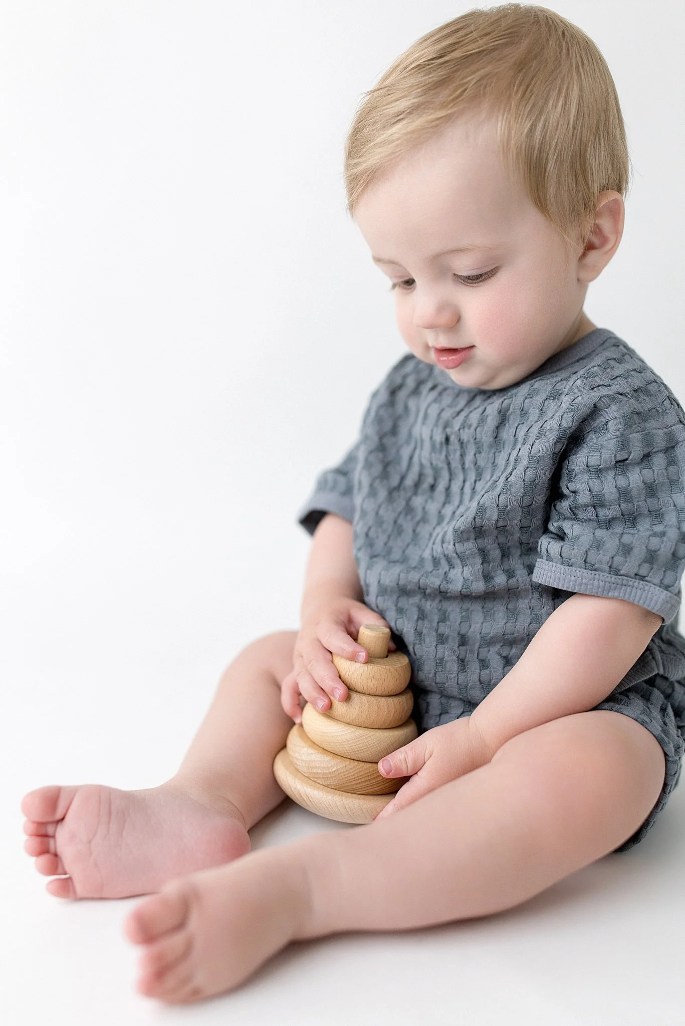 A baby with light skin and blonde hair, sitting on a white surface, holding a wooden stacking toy with rings of different sizes, wearing a grey textured shirt for infant photographers near me.