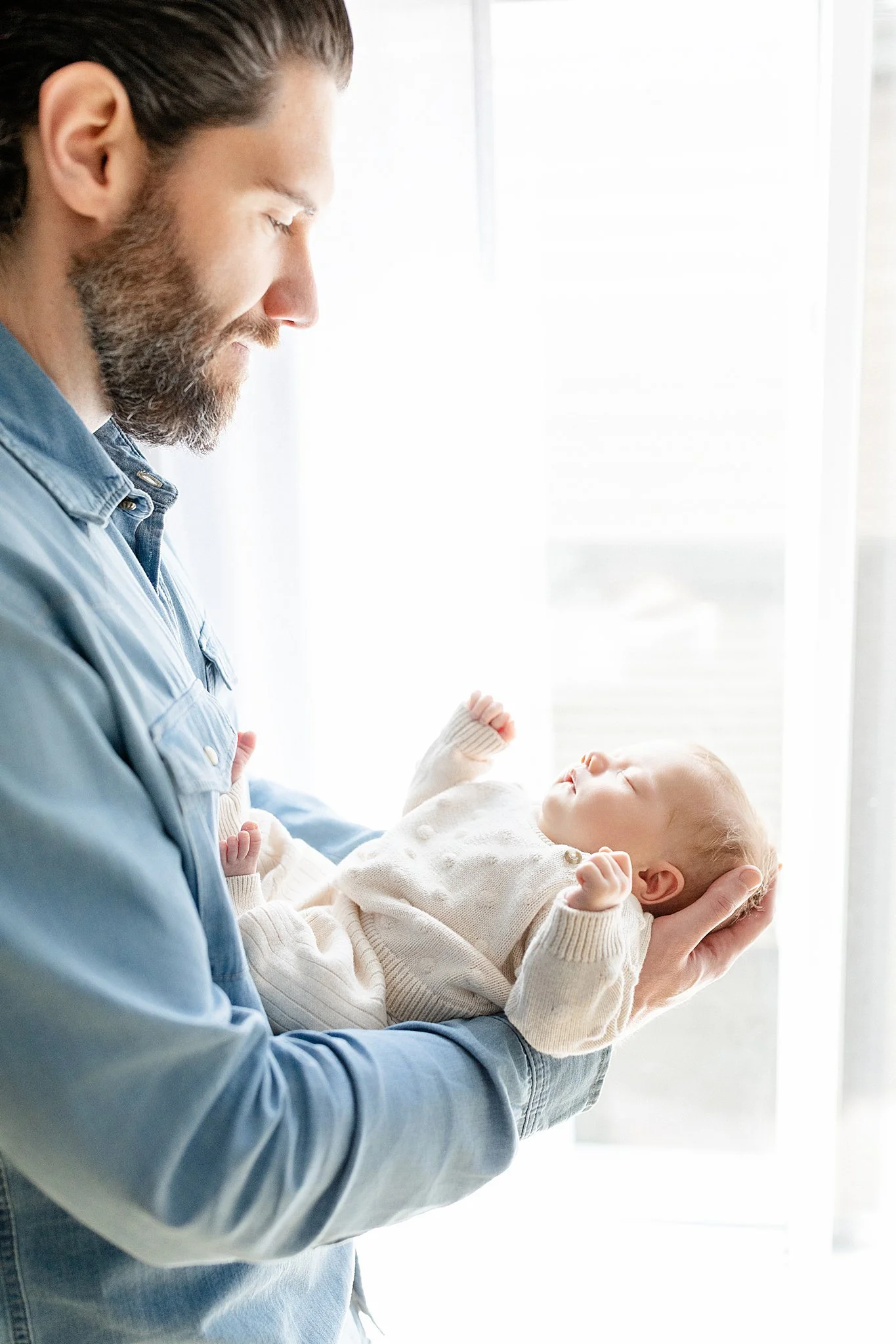 A man with a beard and long hair holding a sleeping baby in his arms near a bright window during in home newborn photos near me.