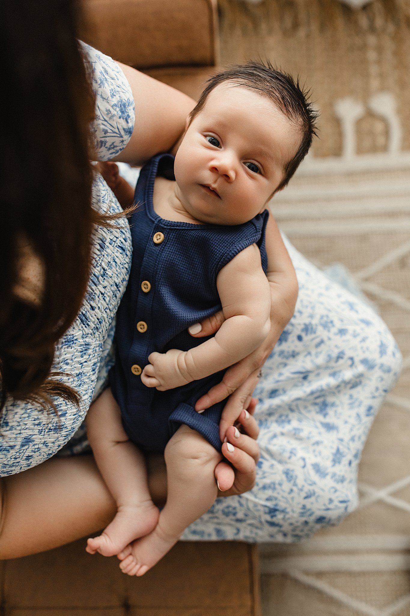 A baby being cradled in an adult's arms, lying on a bed with a patterned blue and white sheet, with a beige headboard and wall with beige and brown textures in the background with Marie Lopez photography.