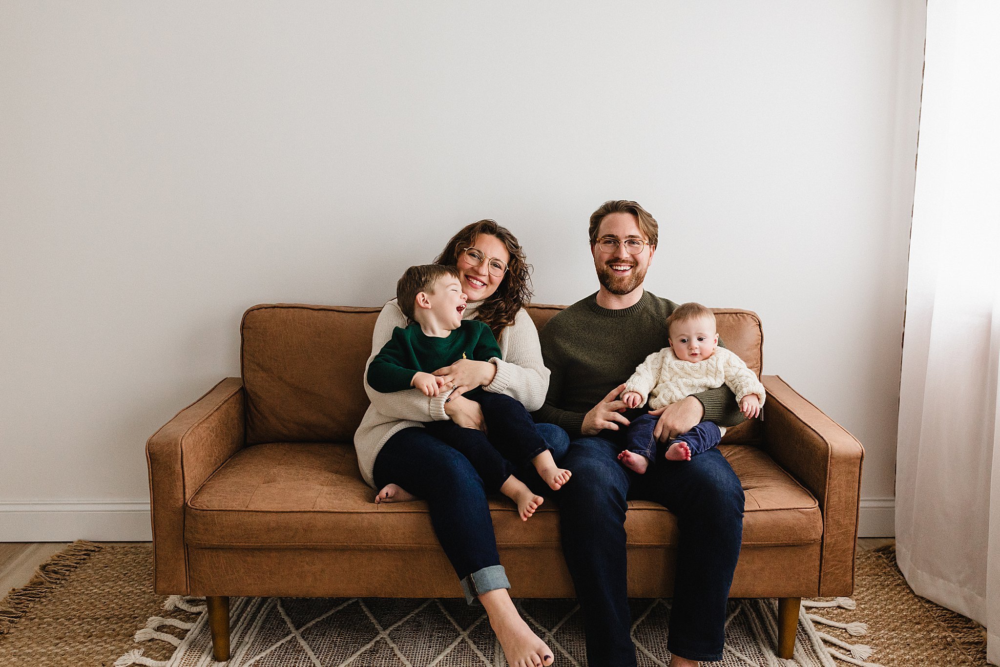 A smiling family of four sitting on a brown couch in a minimally decorated room, with two children and two adults, against a plain white wall and a window with curtains on the right during a studio family photo session in Naperville.