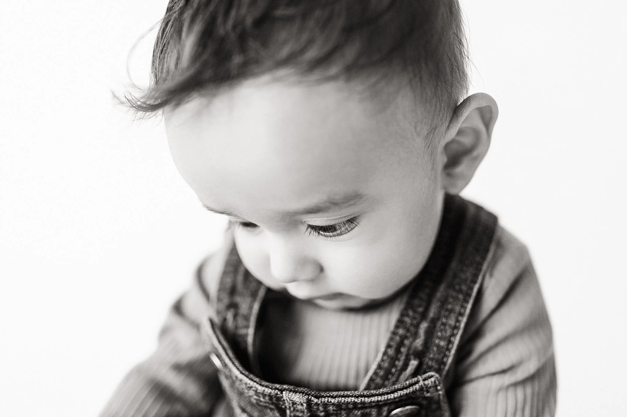Black and white photo of a young child looking downward, with short hair and wearing overalls and a striped shirt during a baby portrait session in Naperville, IL.