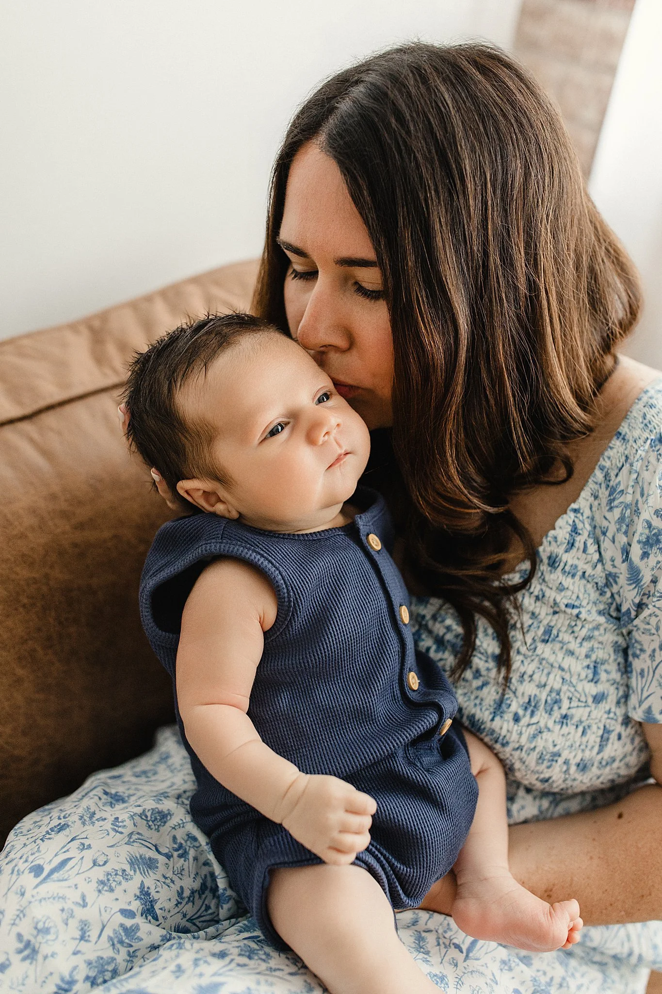 A woman with brown hair and a patterned blue blouse is holding and kissing a baby boy who is wearing a navy blue sleeveless outfit with buttons, sitting on her lap, with a neutral background with newborn photographers near Naperville.