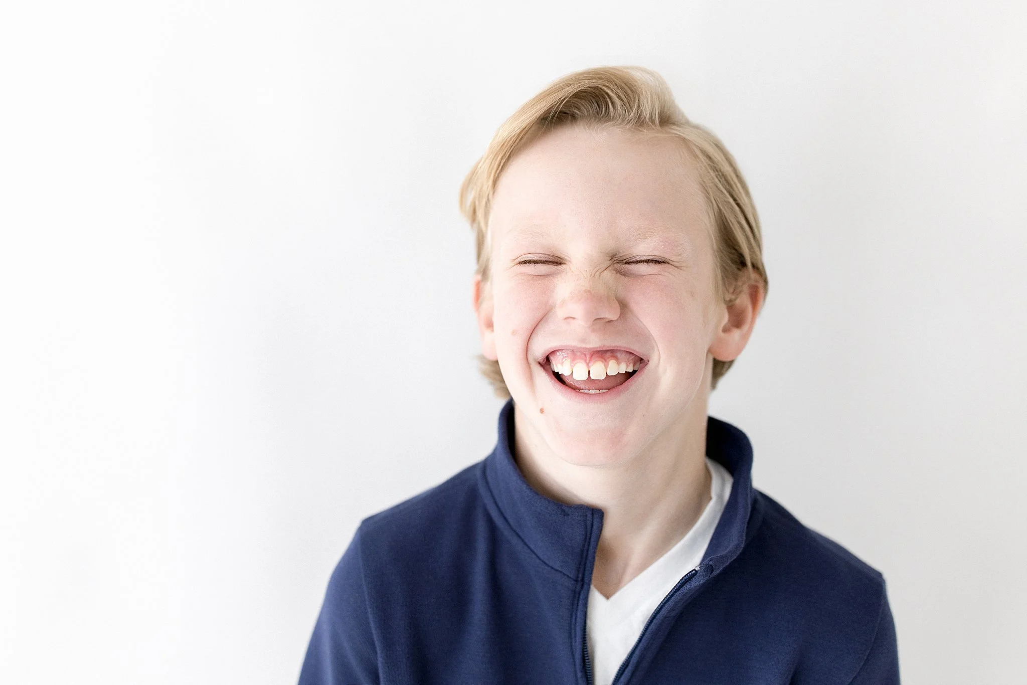 A young boy with blonde hair smiling with eyes closed, wearing a navy blue jacket and a white shirt, standing against a plain white background for comp card photos in Naperville, IL.