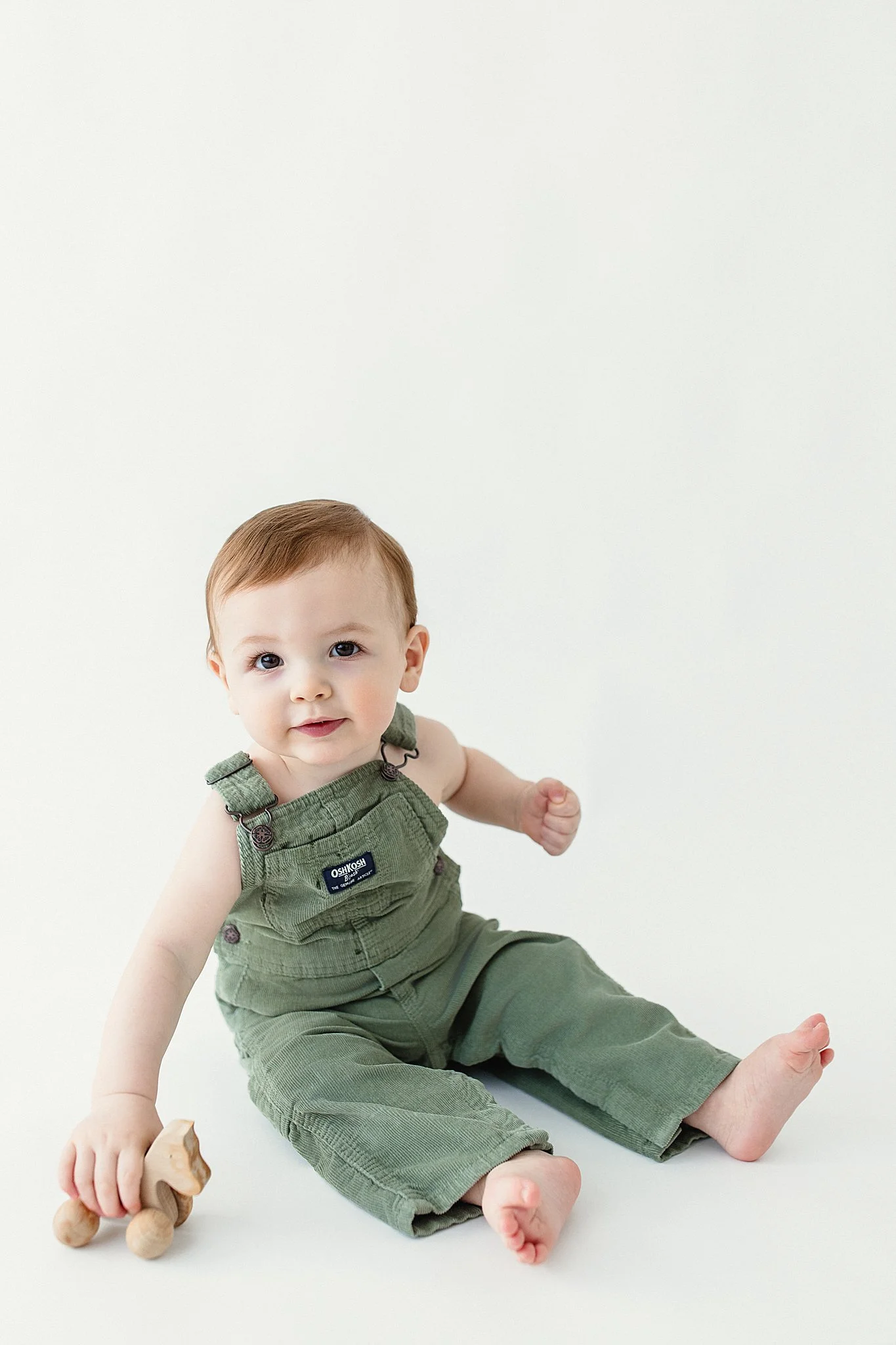 A toddler with brown hair, wearing green overalls, sitting on the floor with a wooden toy in hand, against a plain white background during one year photos near me.