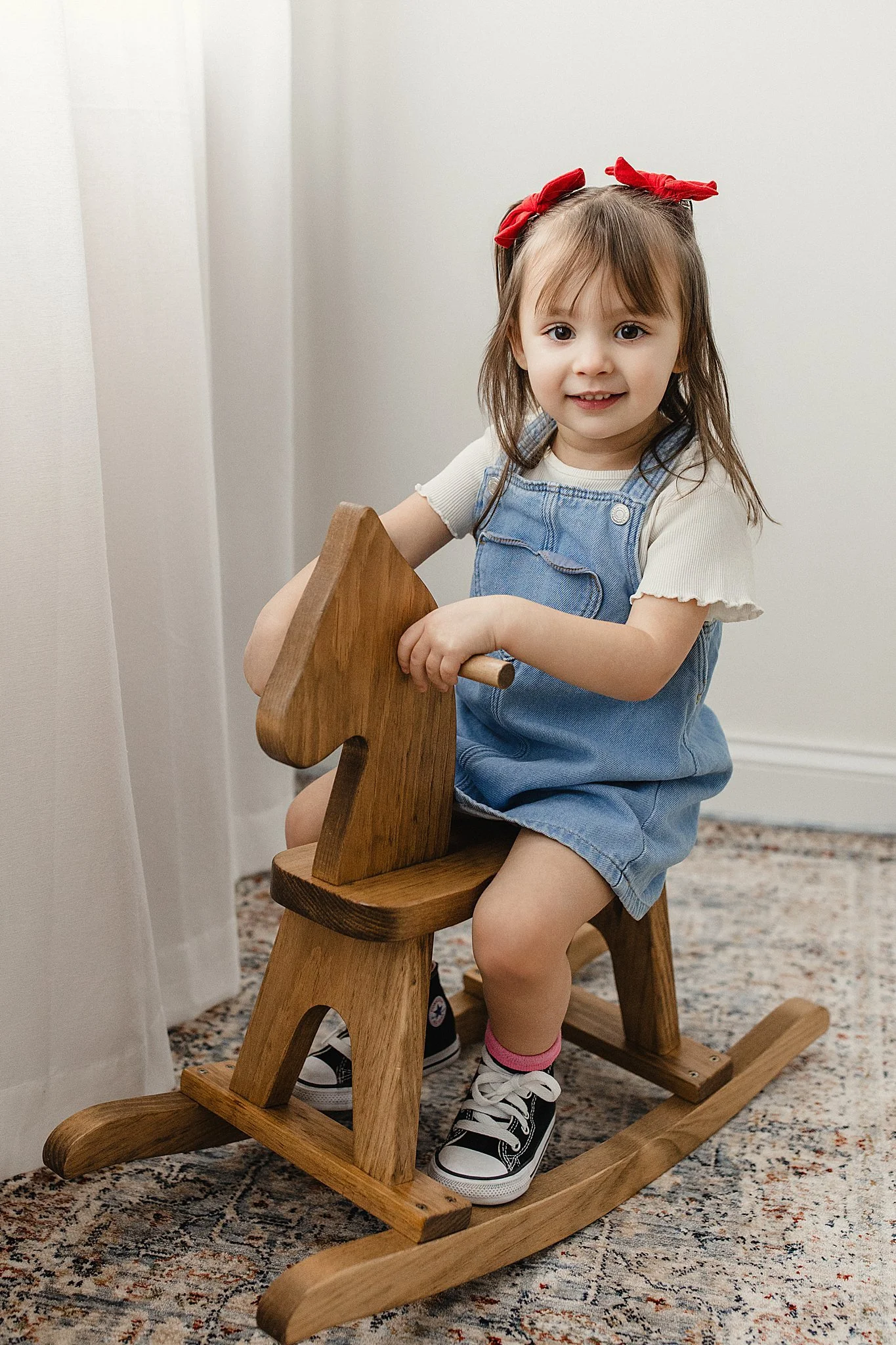 A young girl with brown hair, red hair bows, wearing a white t-shirt, blue overalls, and sneakers, sitting on a wooden rocking horse indoors during a kids photoshoot in downtown Naperville, IL.