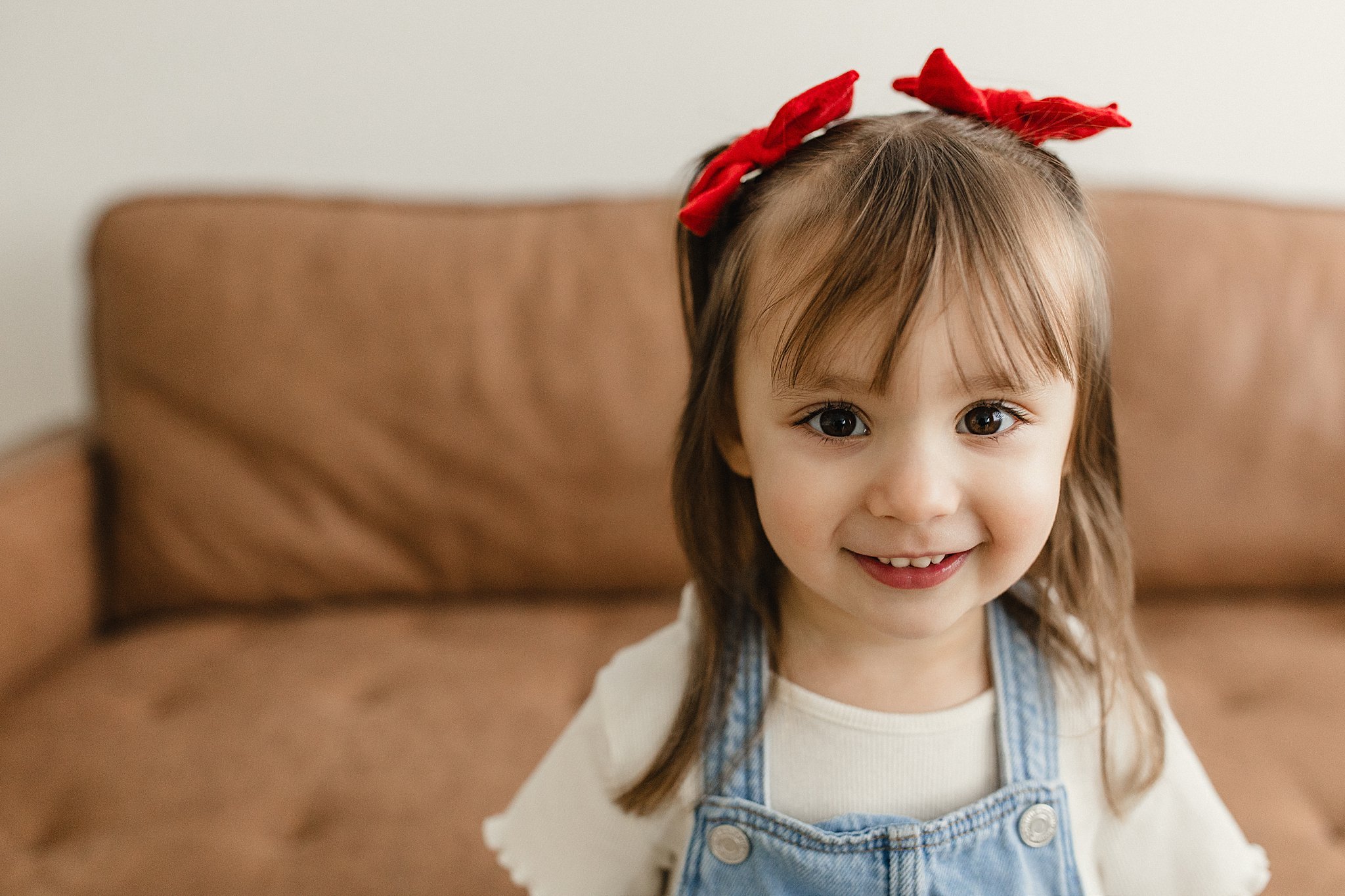 A young girl with light brown hair, wearing a white shirt, denim overalls, and a red bow headband, smiling in front of a brown couch for kids photography near me.