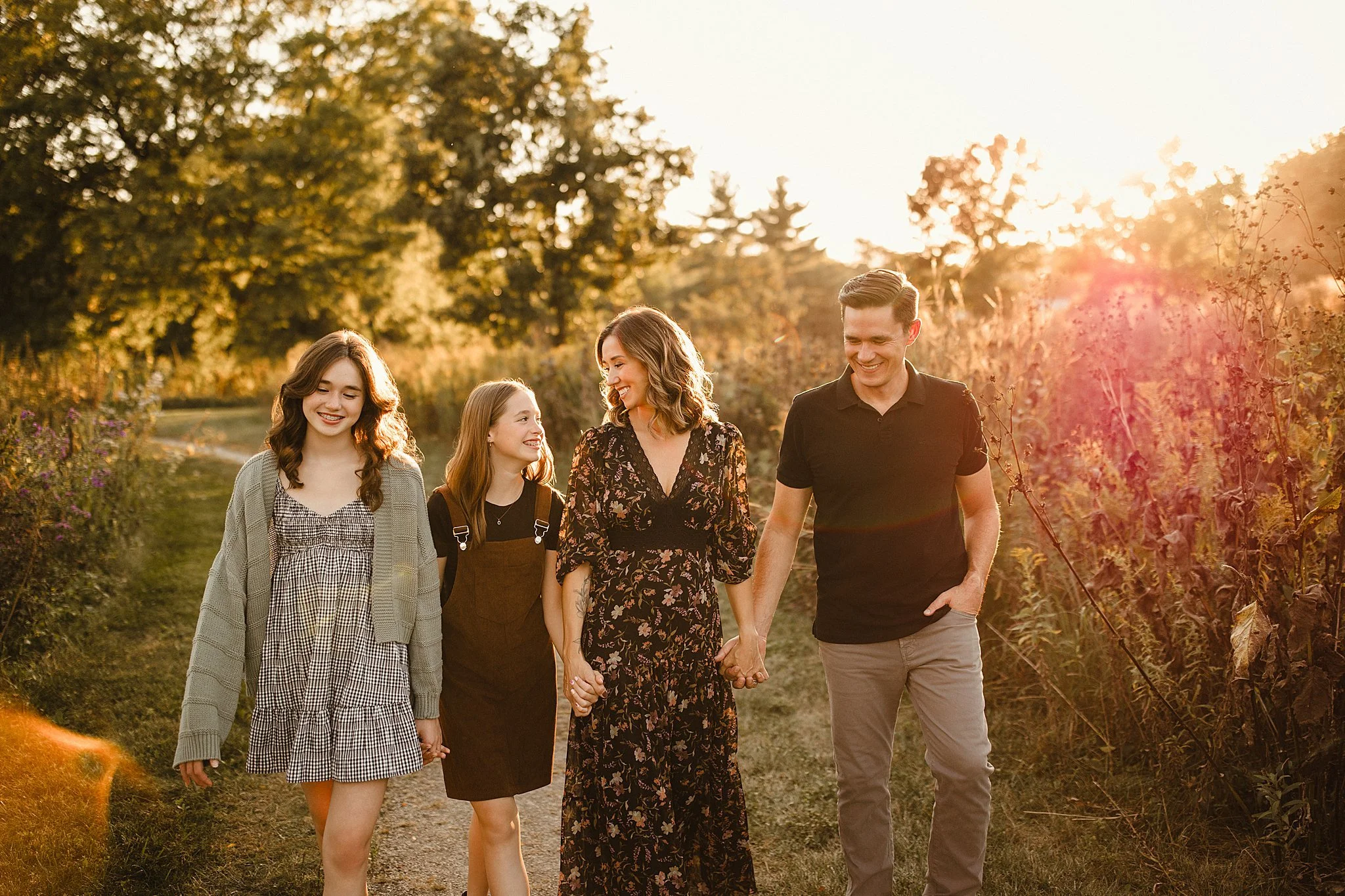 A family of four holding hands and walking through a path in a field during sunset, with trees in the background.