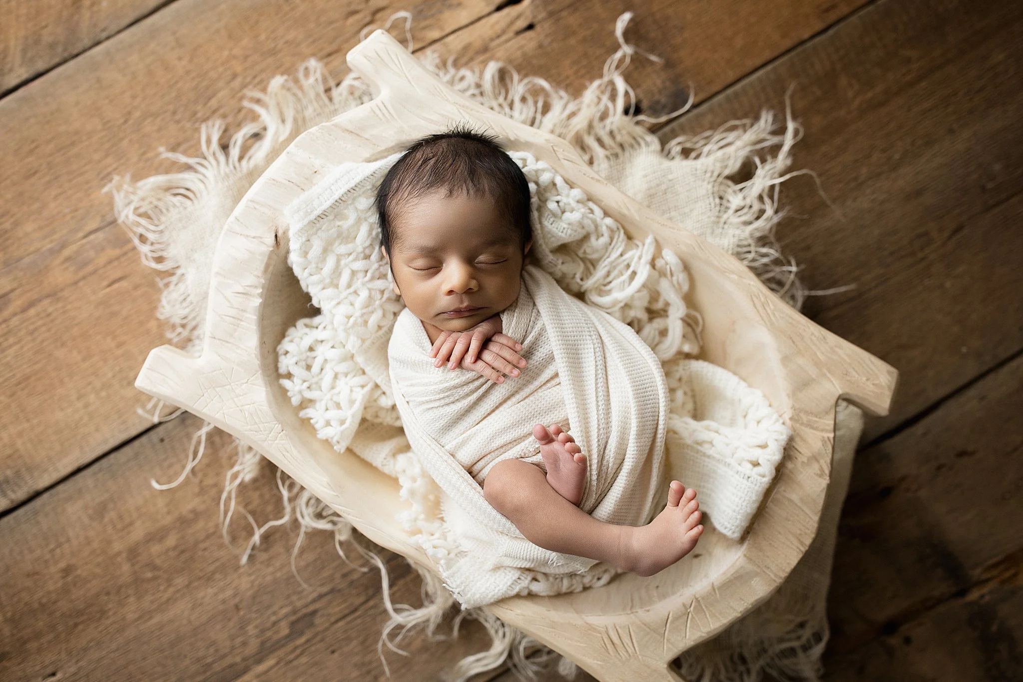 A sleeping baby wrapped in a cream-colored blanket, lying on a wooden cradle with a fluffy white blanket, on a wooden floor with a naperville newborn photographer.