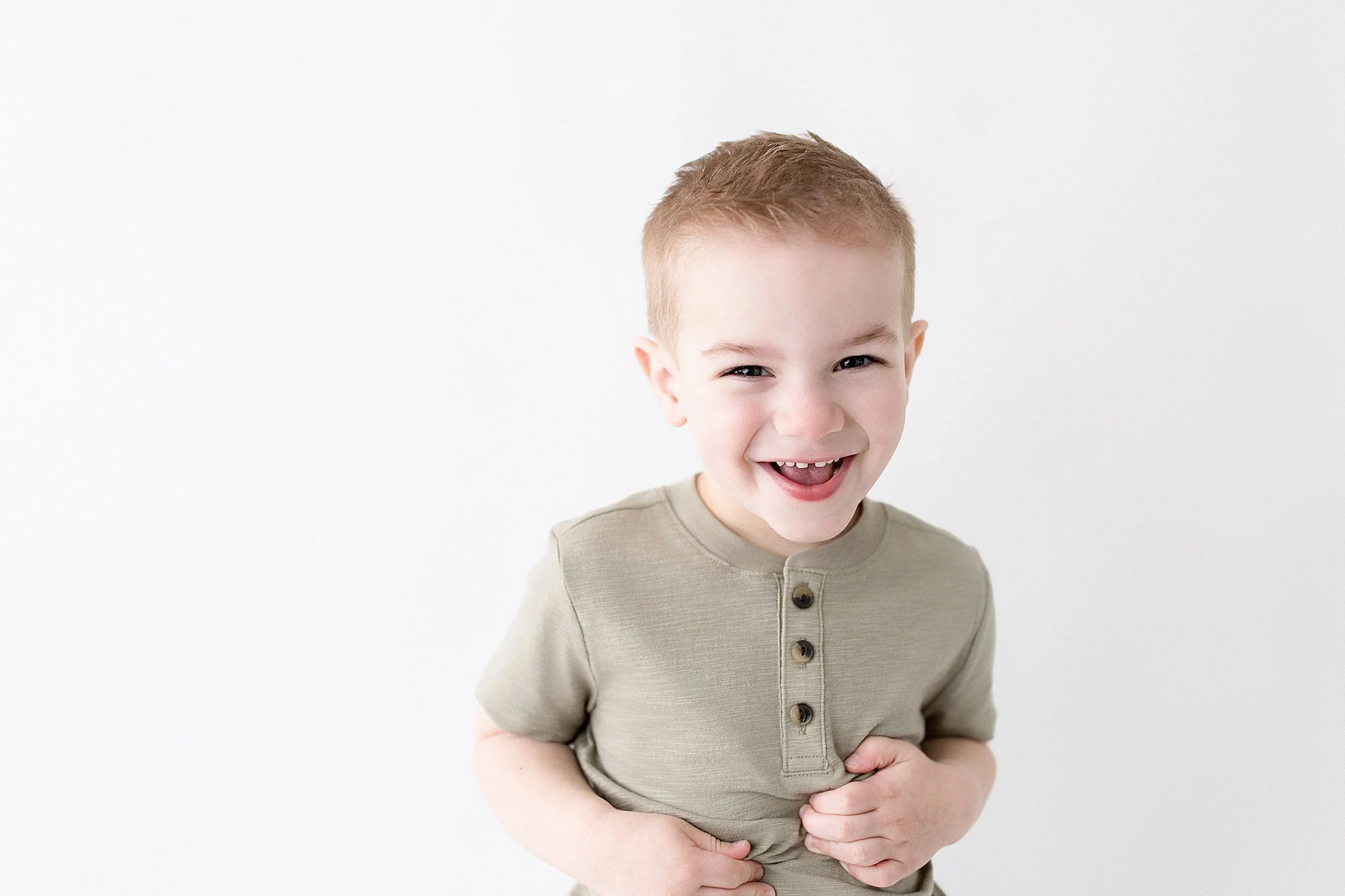 A young boy with short, light brown hair smiling and laughing, wearing a beige shirt with four buttons, standing against a plain white wall with the best children's photographer in Naperville, IL.