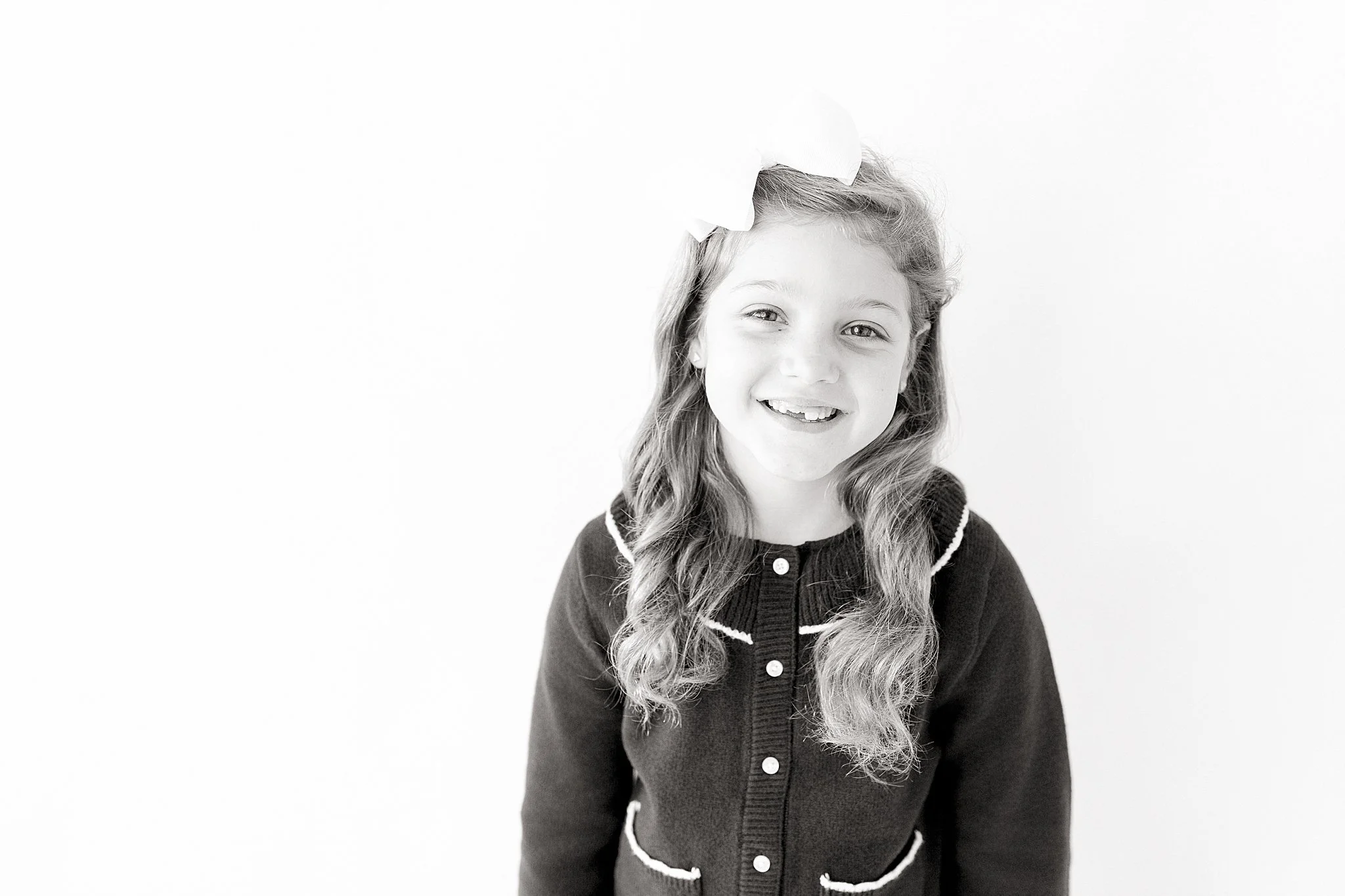 A young girl with curly hair and a big smile, wearing a dark cardigan with white piping and buttons, standing against a plain white background during a personality mini session near me.
