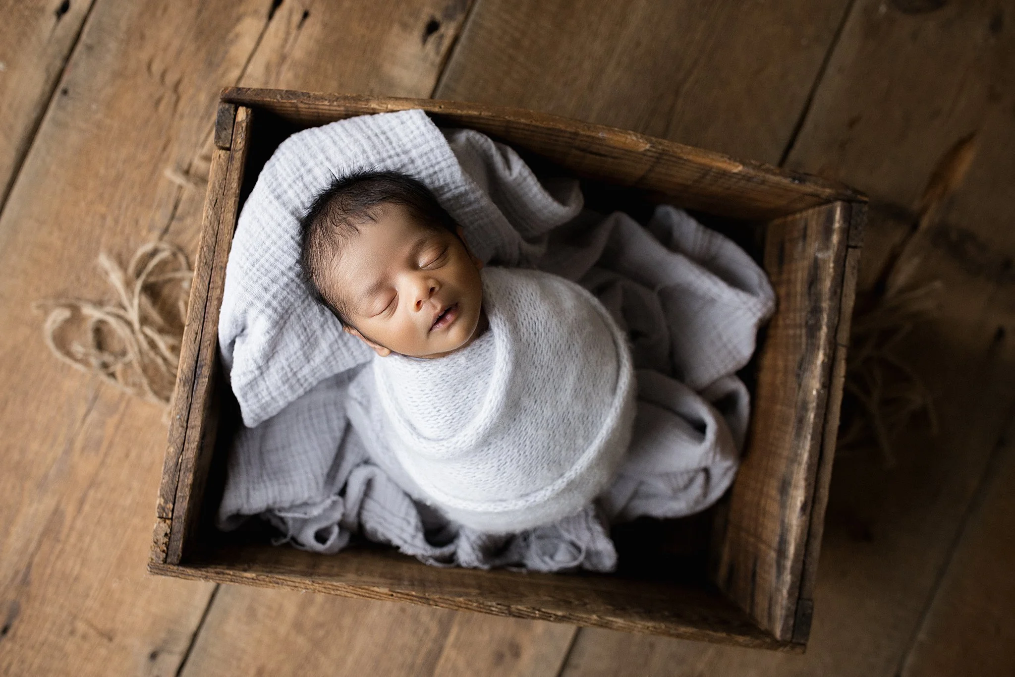 A sleeping newborn baby wrapped in white cloth, lying in a wooden box with a blanket, on a wooden floor with the best newborn photographer in Naperville.