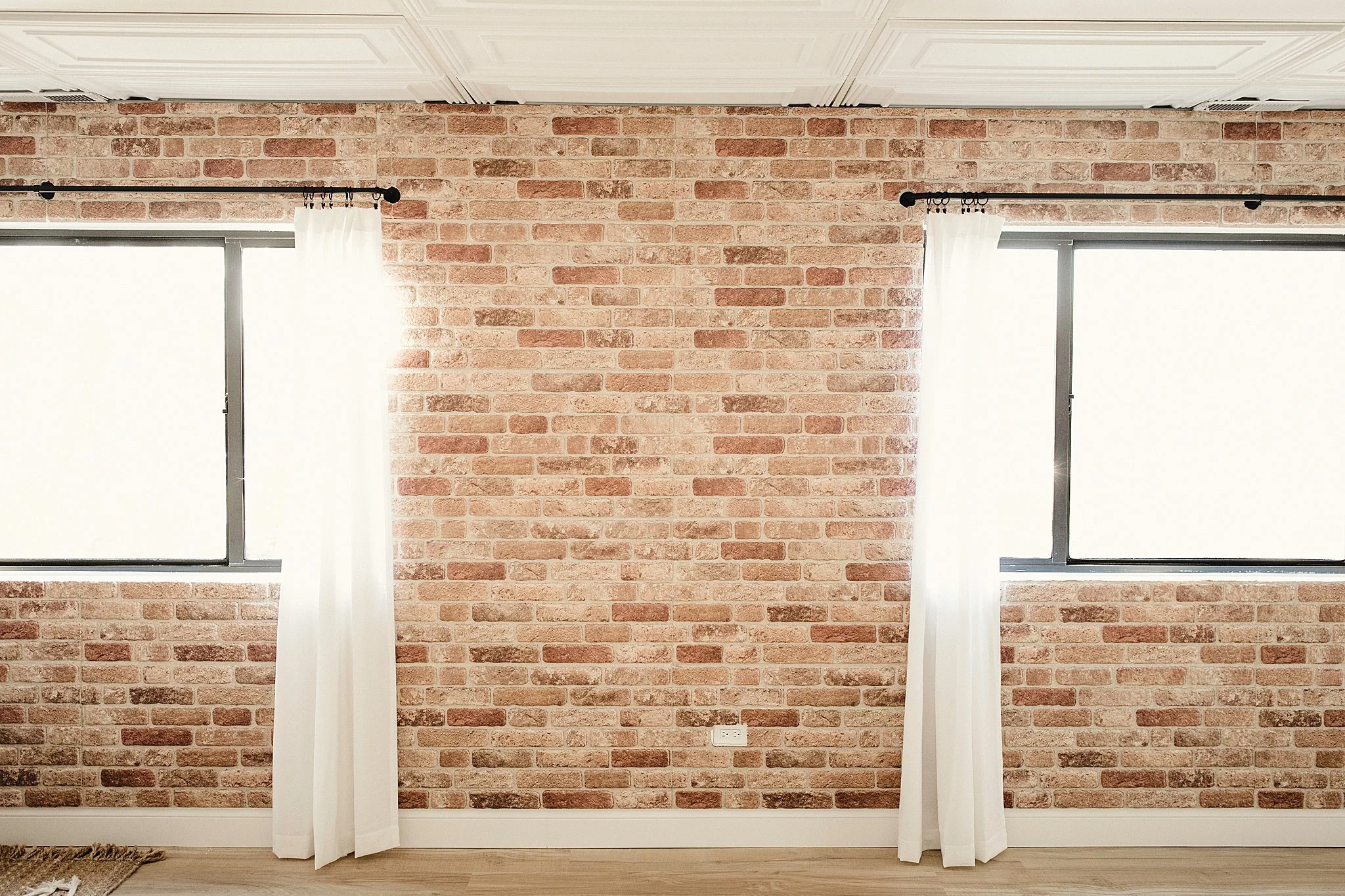 Bright room with a brick wall, two large windows with sheer white curtains, and a white electrical outlet in photo studios near me.