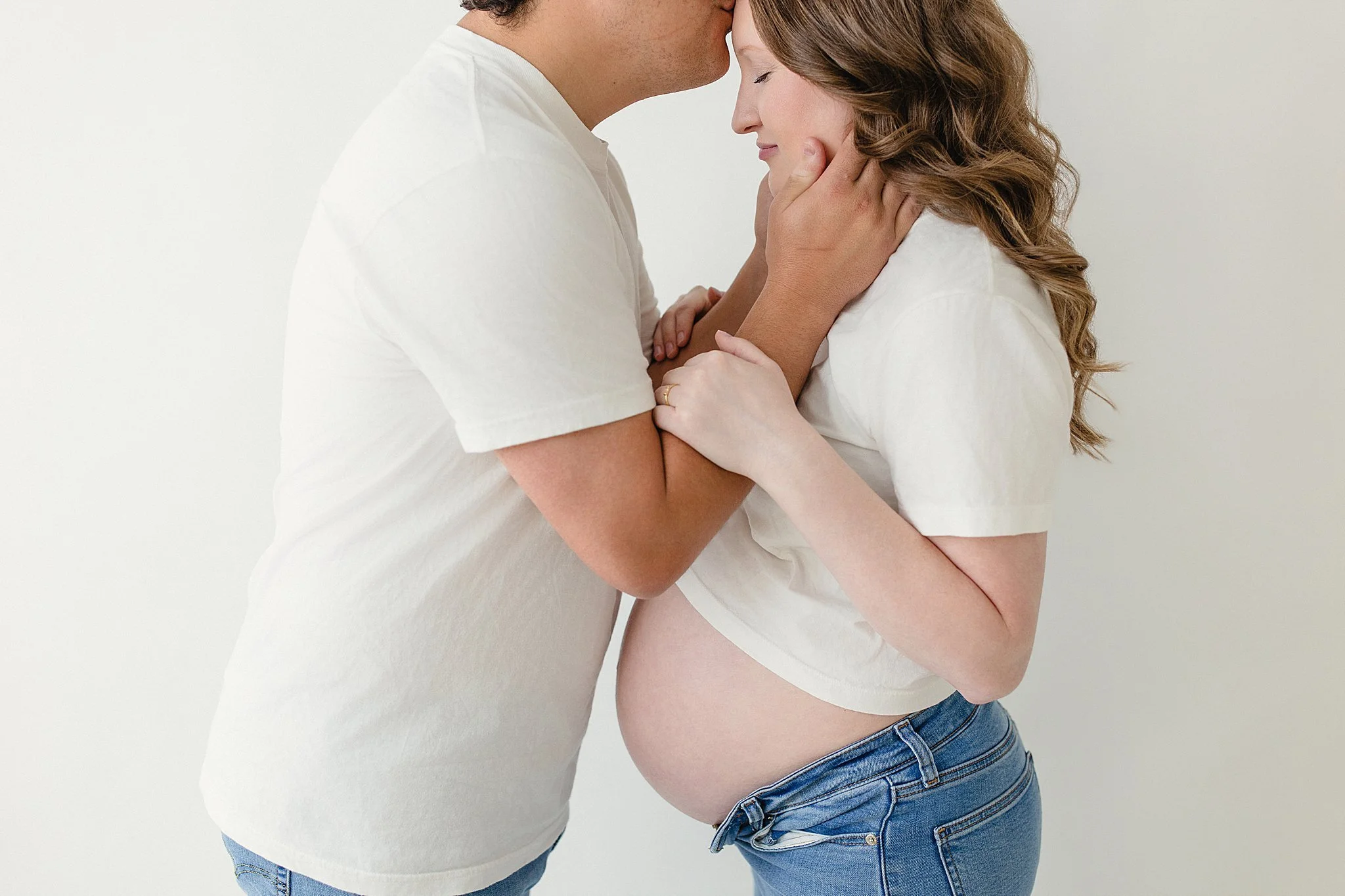 A pregnant woman and her partner sharing a tender moment, with the partner gently touching the woman's face and forehead as she leans into him, both dressed in white shirts and blue jeans, standing for studio maternity photos near me.