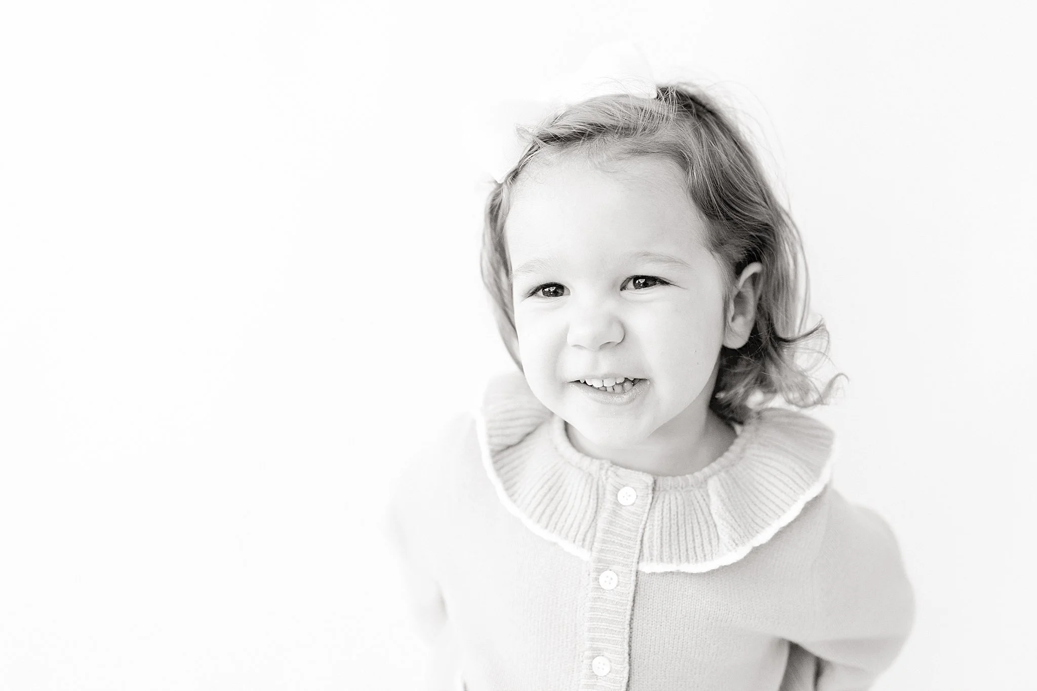 A young girl with wavy hair smiling in front of a plain white background during black and white portraits near me.