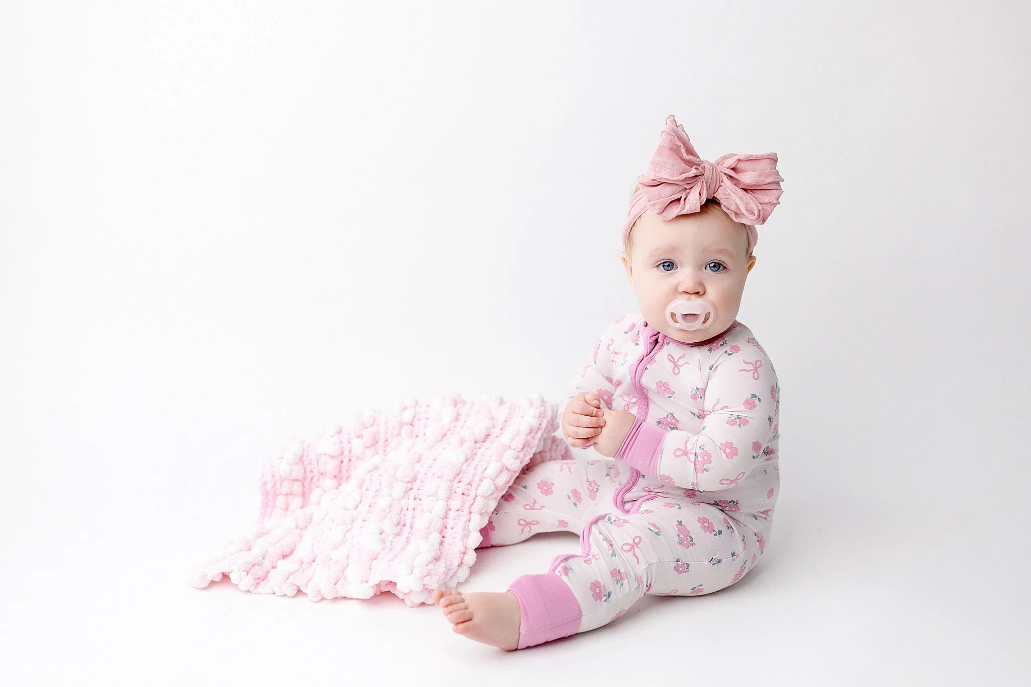 A baby girl sitting on a pink blanket against a white background, wearing pink and white pajamas with a cherry blossom pattern and a pink headscarf tied in a bow, with a pink pacifier in her mouth with Ally and B Photography.