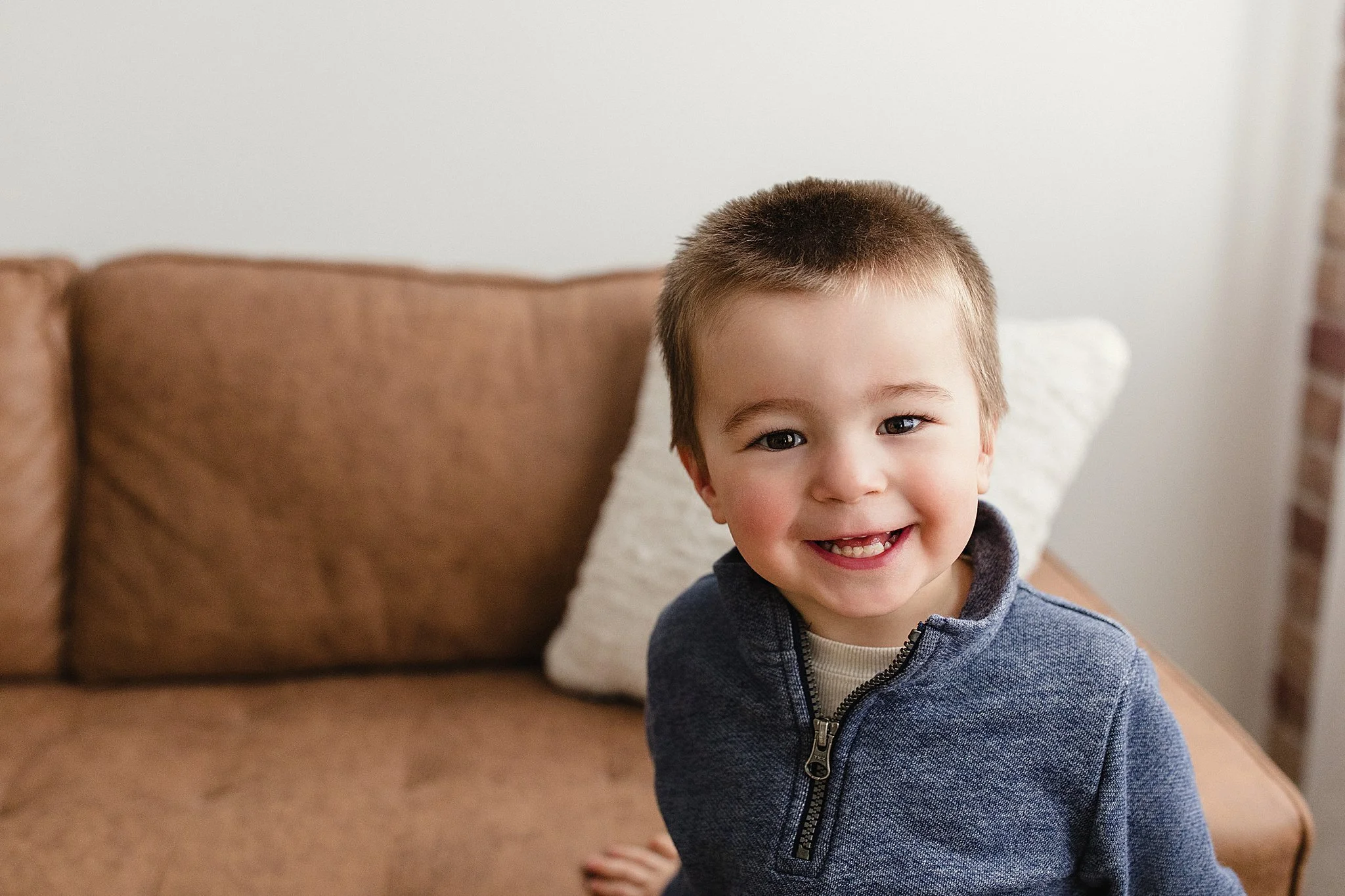 A young boy with short brown hair and a big smile, wearing a blue zip-up sweater, sitting on a brown sofa with a white pillow behind him in a cozy living room with a kids photographer Naperville.