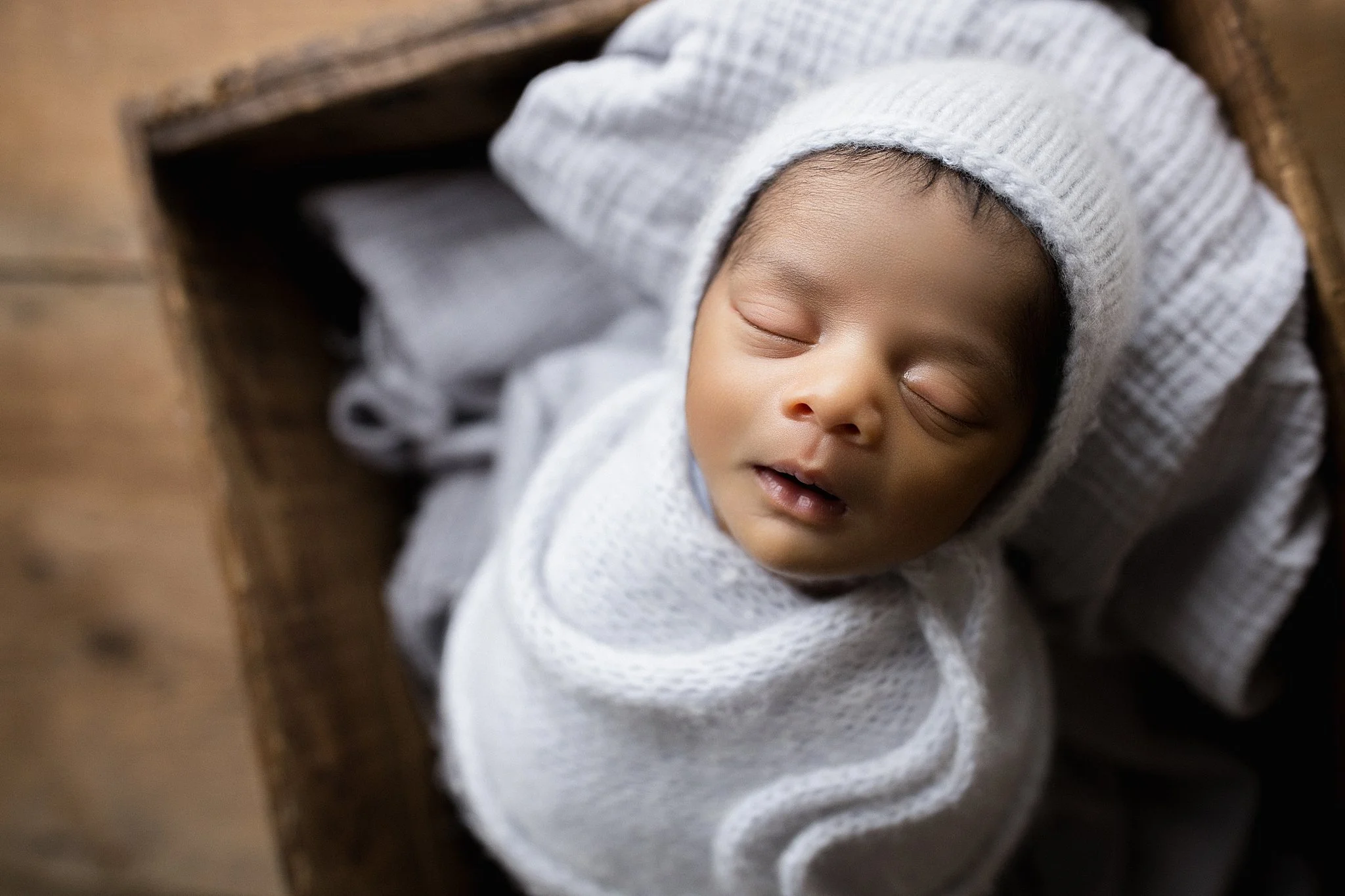 A sleeping baby swaddled in a white blanket, wearing a striped knit hat, lying in a wooden basket for best newborn photos near me.