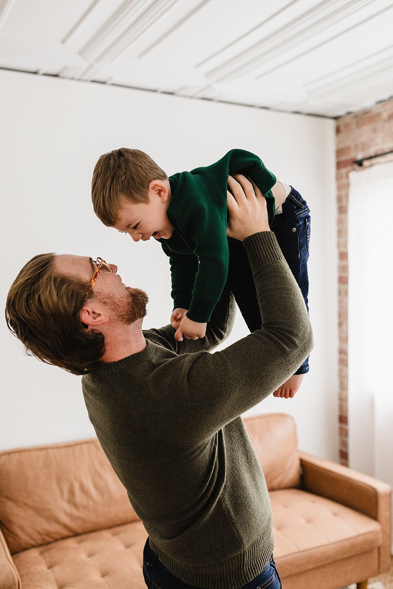 A man with glasses and a beard lifts a young boy in the air while they both smile and laugh inside a living room with a brown couch and a white wall for a Naperville studio family photo sesison.