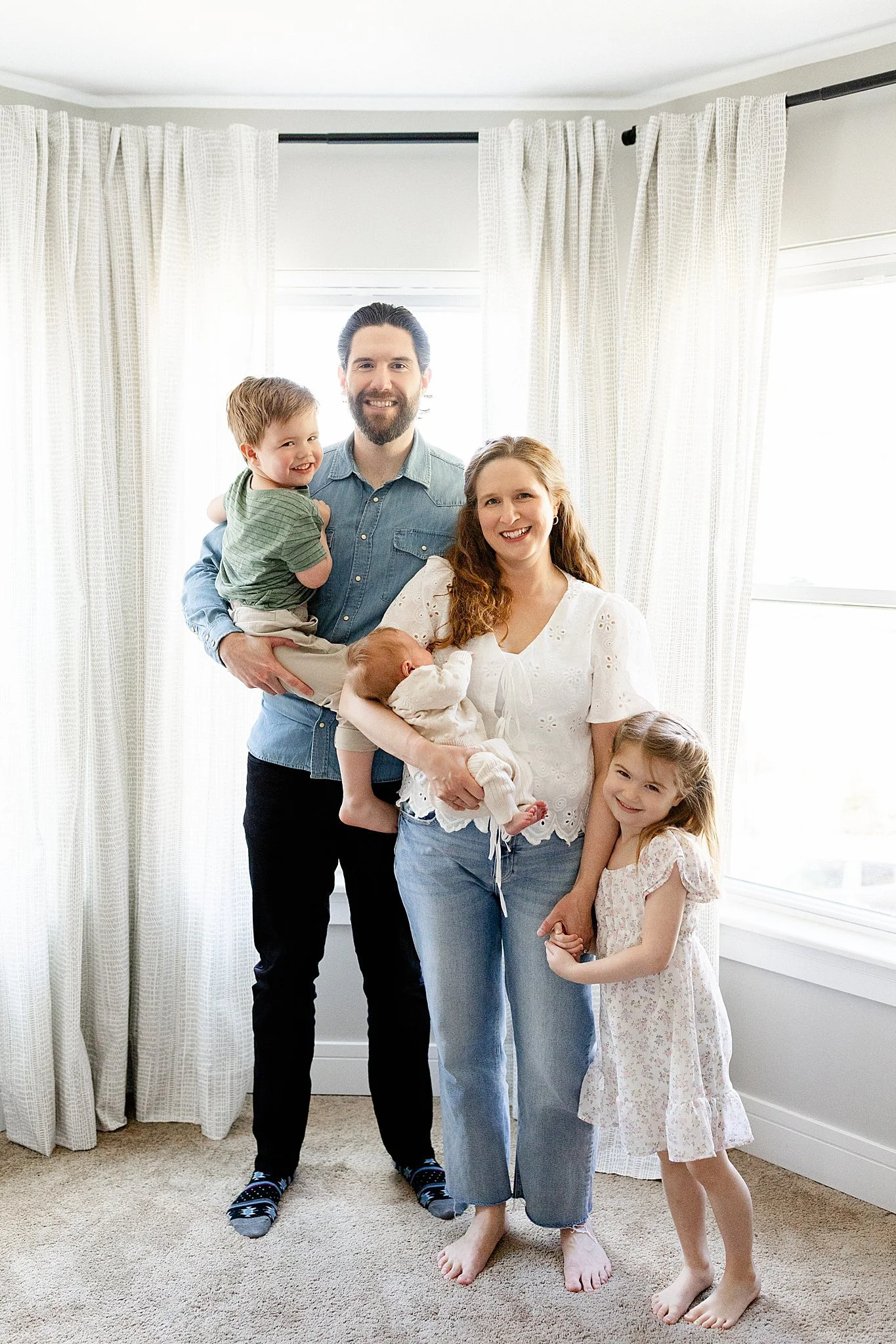 A happy family of five, including a father, mother, and three children, standing inside a bright room near a window with white curtains. The father is holding a young boy, and the mother is holding a sleeping baby while the girl stands beside her.