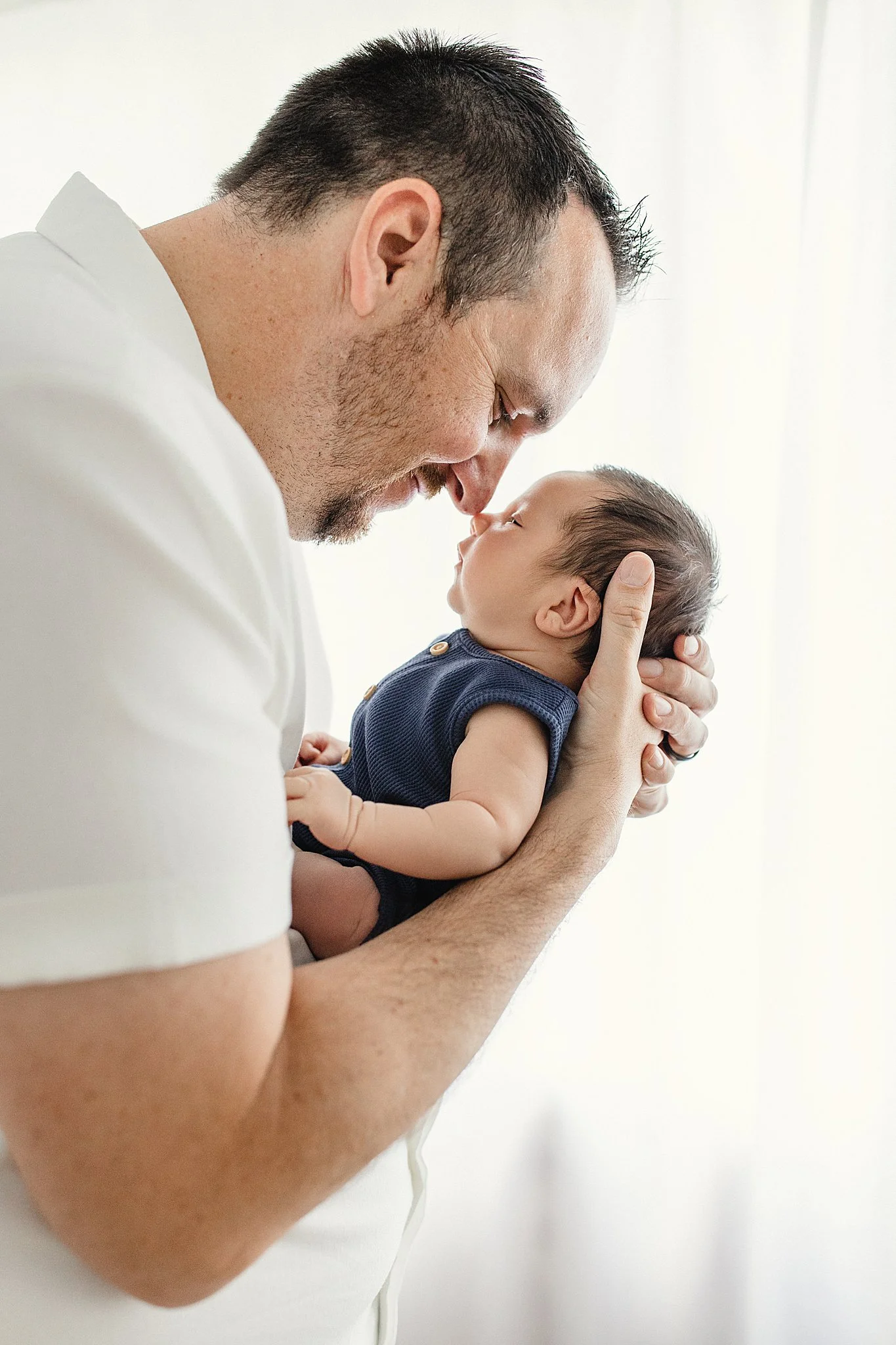 A man holding a baby close with their foreheads touching, both with eyes closed in a tender moment during lifestyle newborn photos near me.