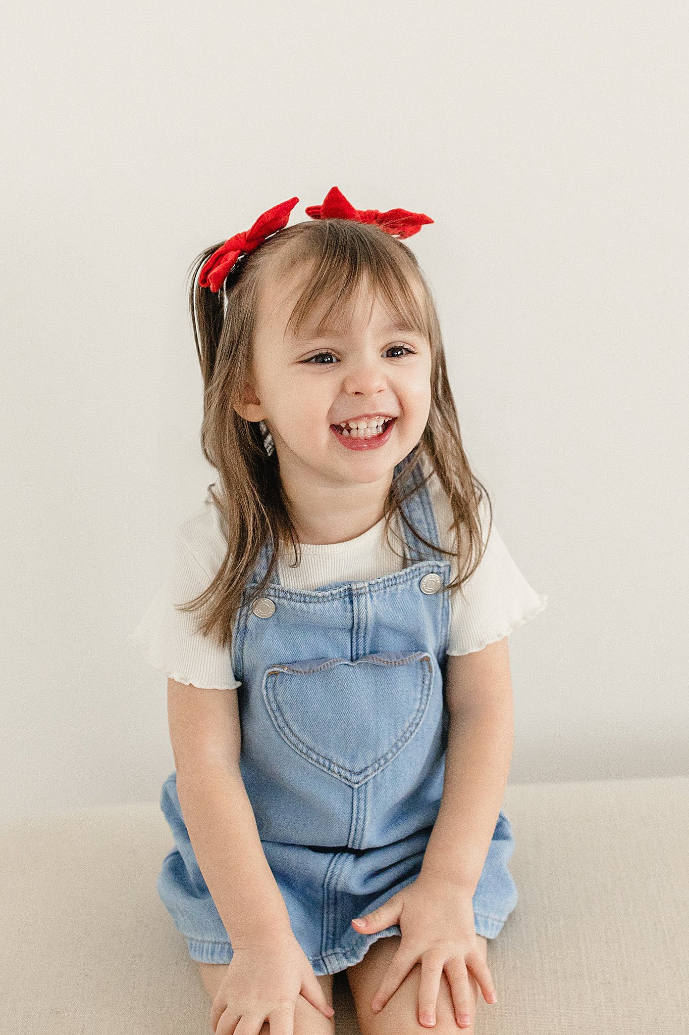 A smiling young girl with light brown hair, wearing a red hair bow and blue denim overalls over a white shirt, sitting against a plain light-colored background with Ally and B Photography.