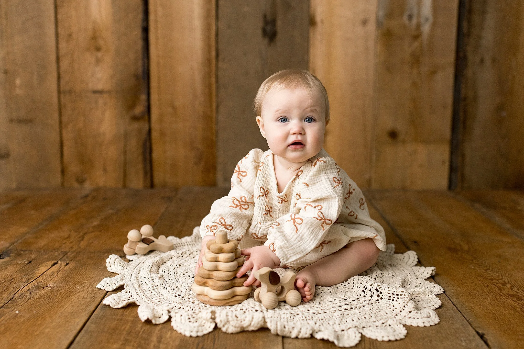 A young baby with light hair and blue eyes, wearing a cream-colored outfit with orange bows, is sitting on a white crochet blanket on a wooden floor. The baby is holding a wooden ring toy and sitting in front of a wooden background during photos.