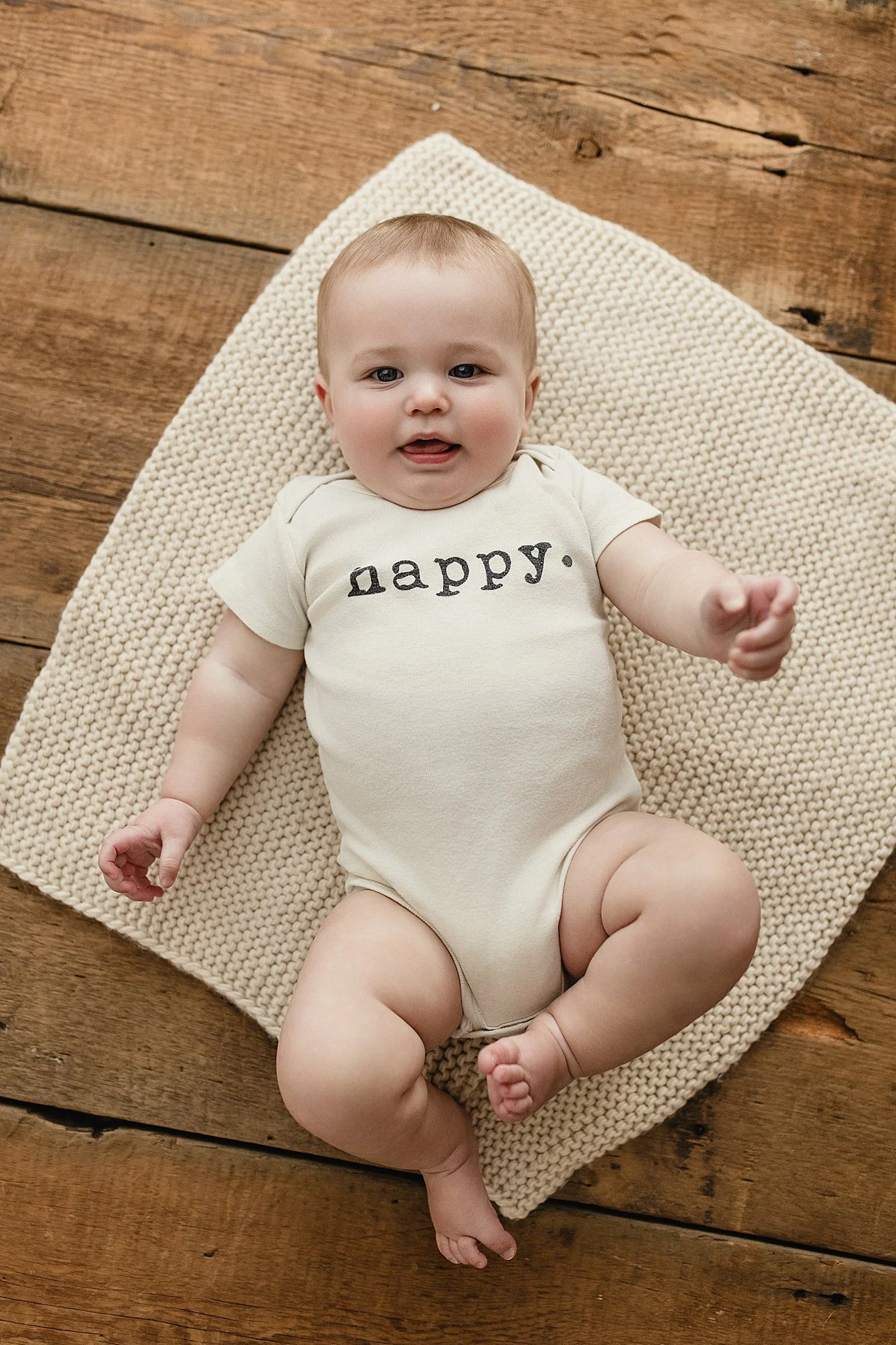 A baby lying on a knit blanket on a wooden floor, wearing a cream-colored onesie with the word 'happy.' printed on it, smiling and reaching out with Naperville baby photographers.