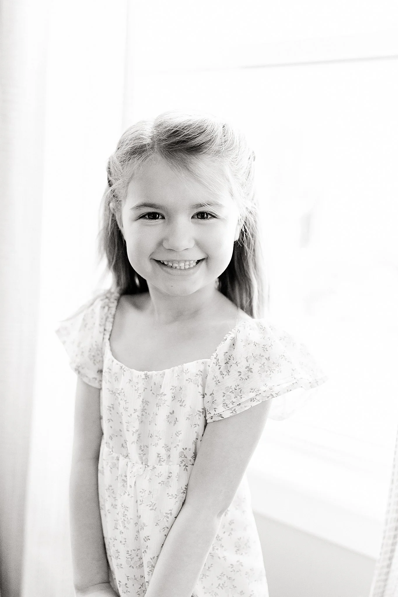 A smiling young girl with long hair, wearing a light-colored dress with a floral pattern, posing indoors near a window during lifestyle photos near me.