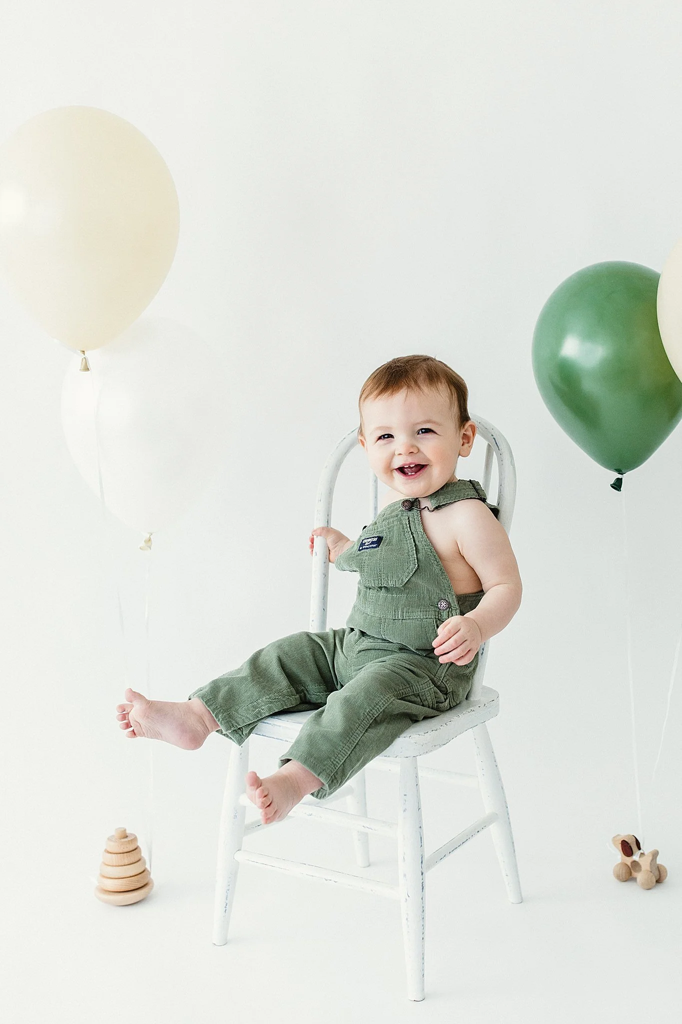 Smiling baby sitting on a white chair surrounded by balloons and toys, celebrating a special occasion with best baby photographer in Naperville, IL.