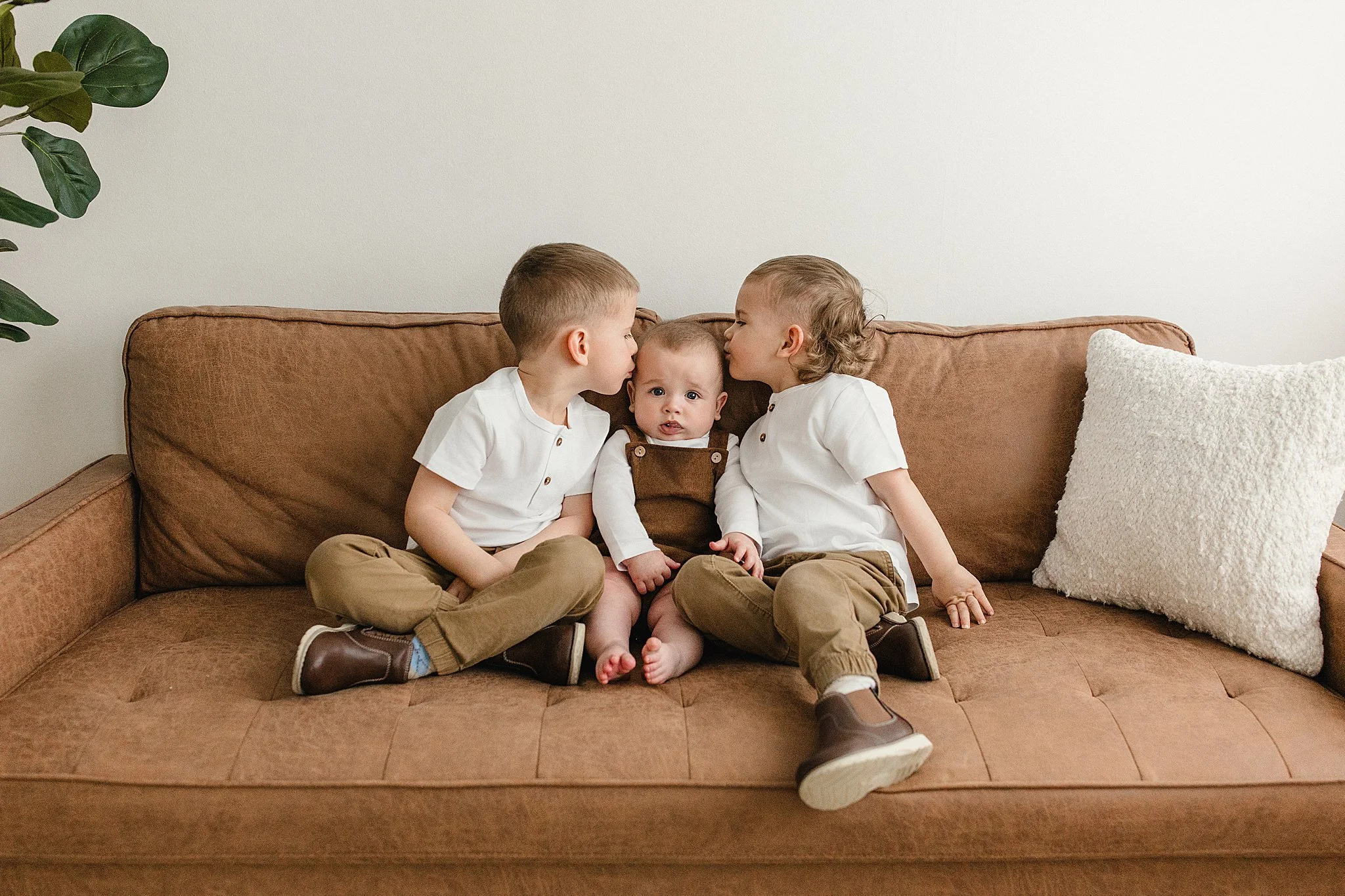 Three young children sitting on a brown couch, with two boys giving kisses to a baby in the middle. All three are dressed in white shirts and tan pants, inside a room with a minimal decor and a white pillow on the right during lifestyle photos.