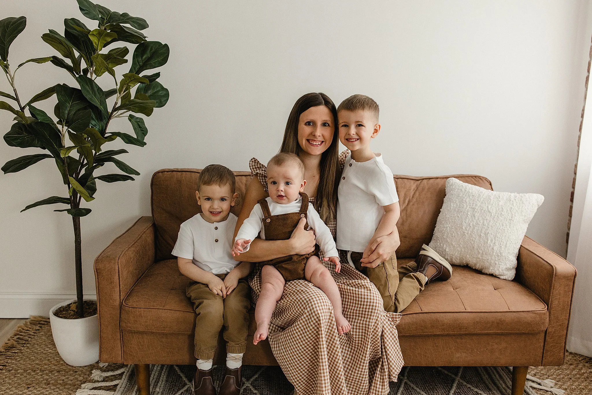 A smiling woman with long brown hair sitting on a brown couch with four young children, all dressed in casual clothing, in a cozy living room with a green potted plant and a white pillow during mommy and me photos with Ally and B Photography.