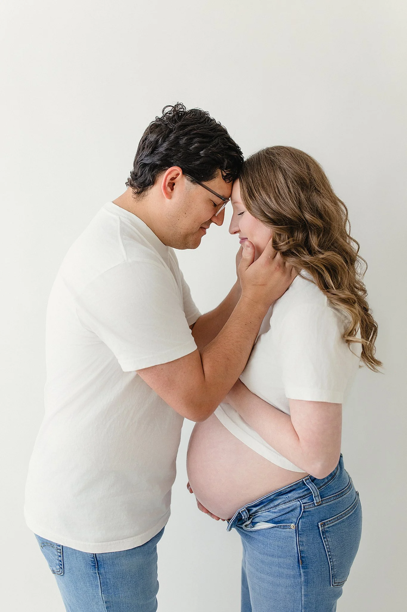 A pregnant woman with long wavy hair and light skin gently touching her belly, with a man with dark hair and glasses touching her forehead gently, both smiling softly against a plain white background during studio maternity session Naperville, IL.