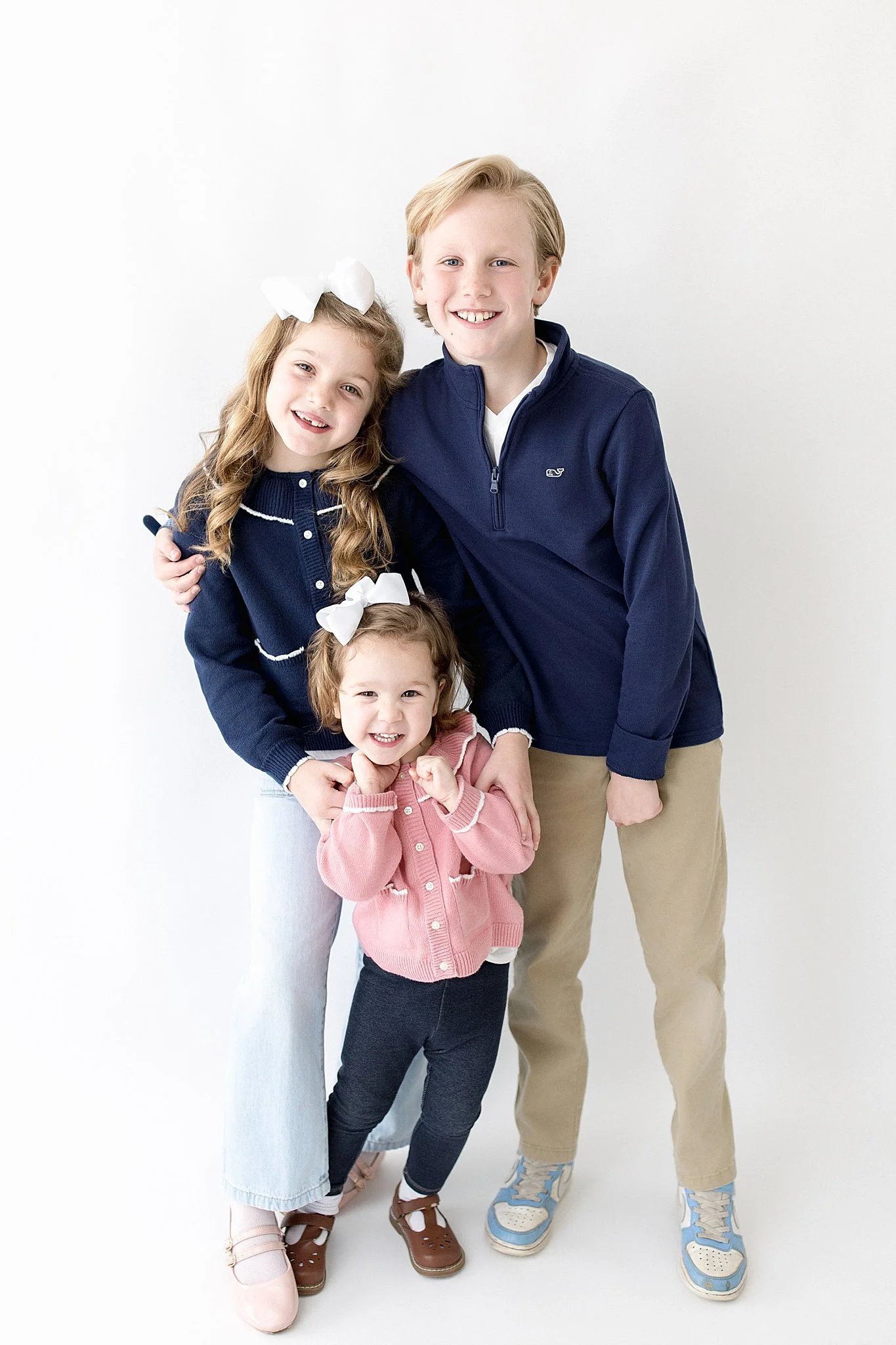 A happy family of four standing together against a white background, including a young girl with a white bow, a younger girl with a white bow, an older girl, and a young man, all smiling and embracing with a kids portrait photographer in Naperville.