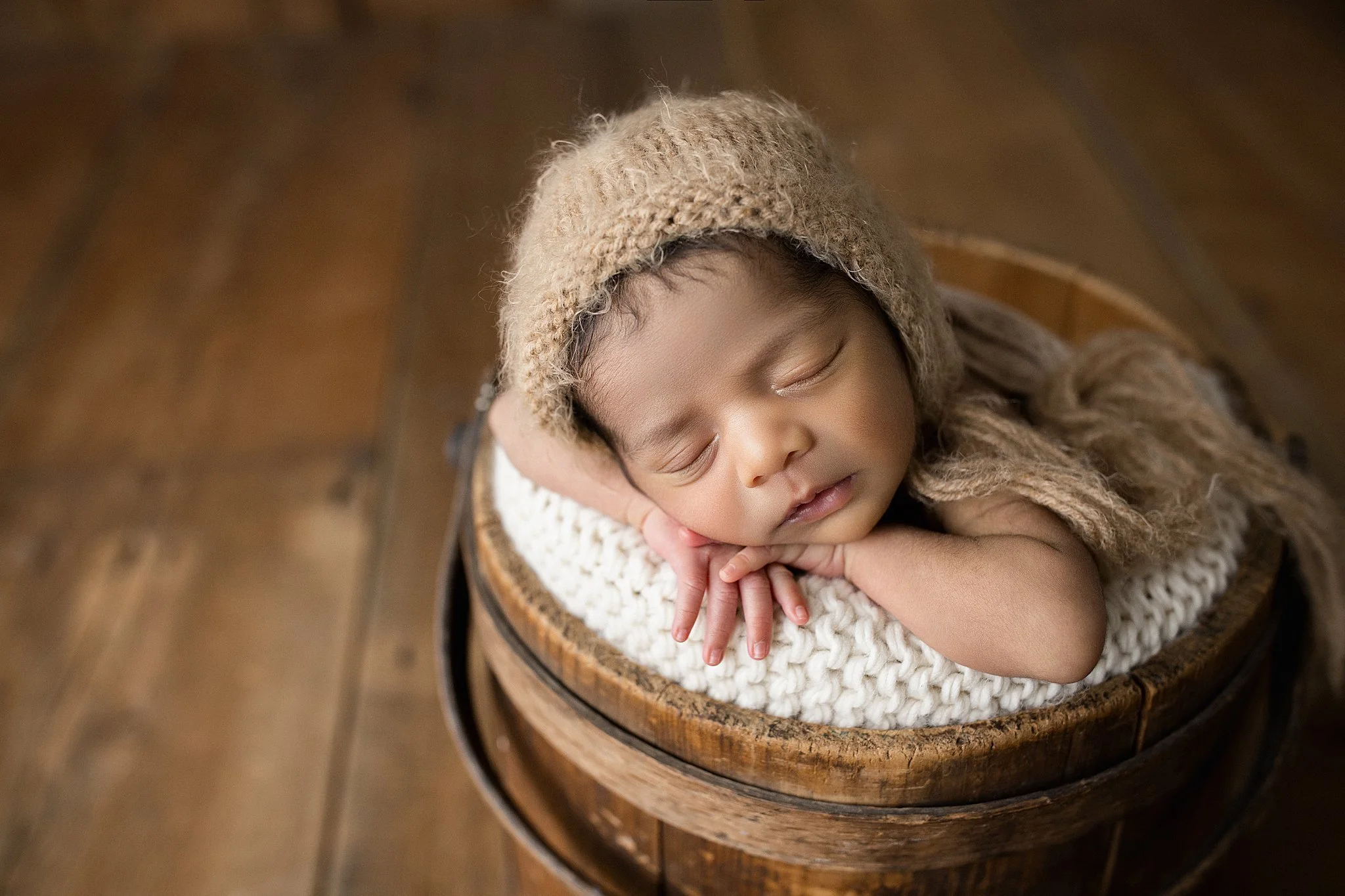A sleeping baby with curly hair, wearing a knitted hat, resting on a soft blanket inside a wooden bucket on a wooden floor during a posed newborn photography photo session.