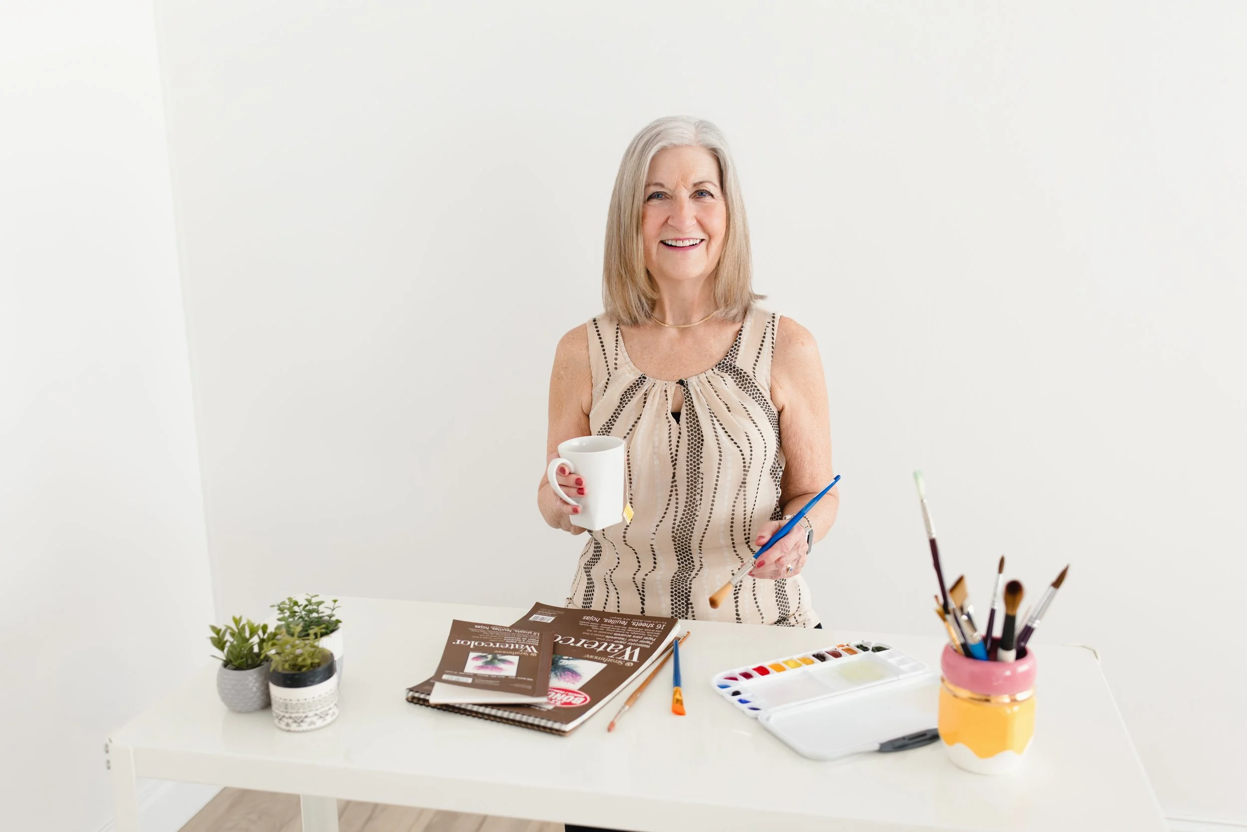 Older woman smiling, holding a mug and a paintbrush, sitting at a white table with art supplies and small potted plants, in a brightly lit room for artist branding photos with Ally and B Photography in Naperville, IL.