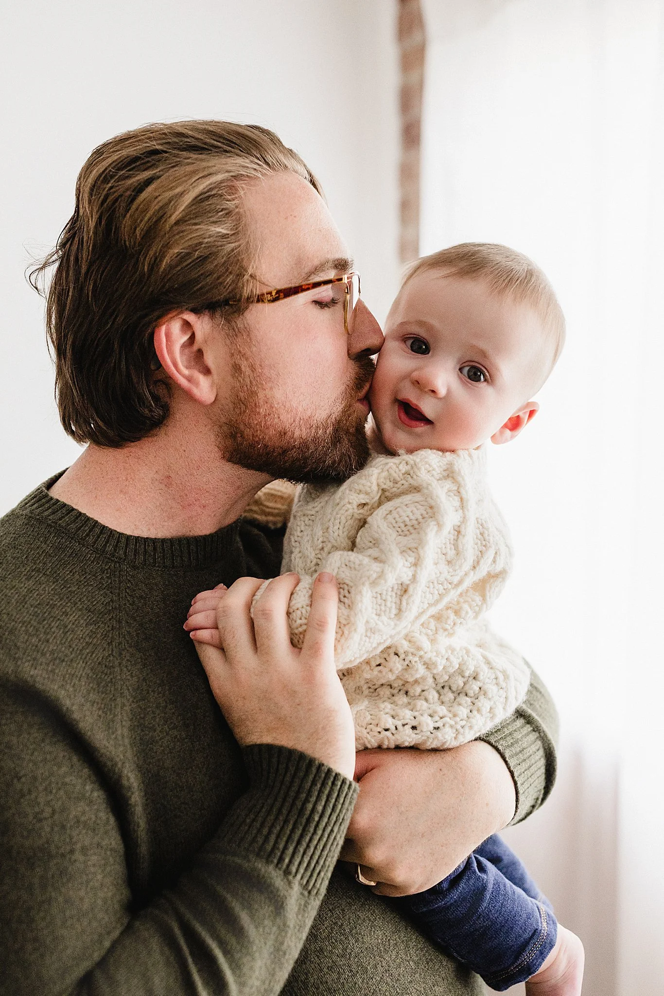 A man with glasses and a beard holding a baby girl, who has light hair and is wearing a cream-colored sweater, close to his face with a family photographer in Naperville, IL.