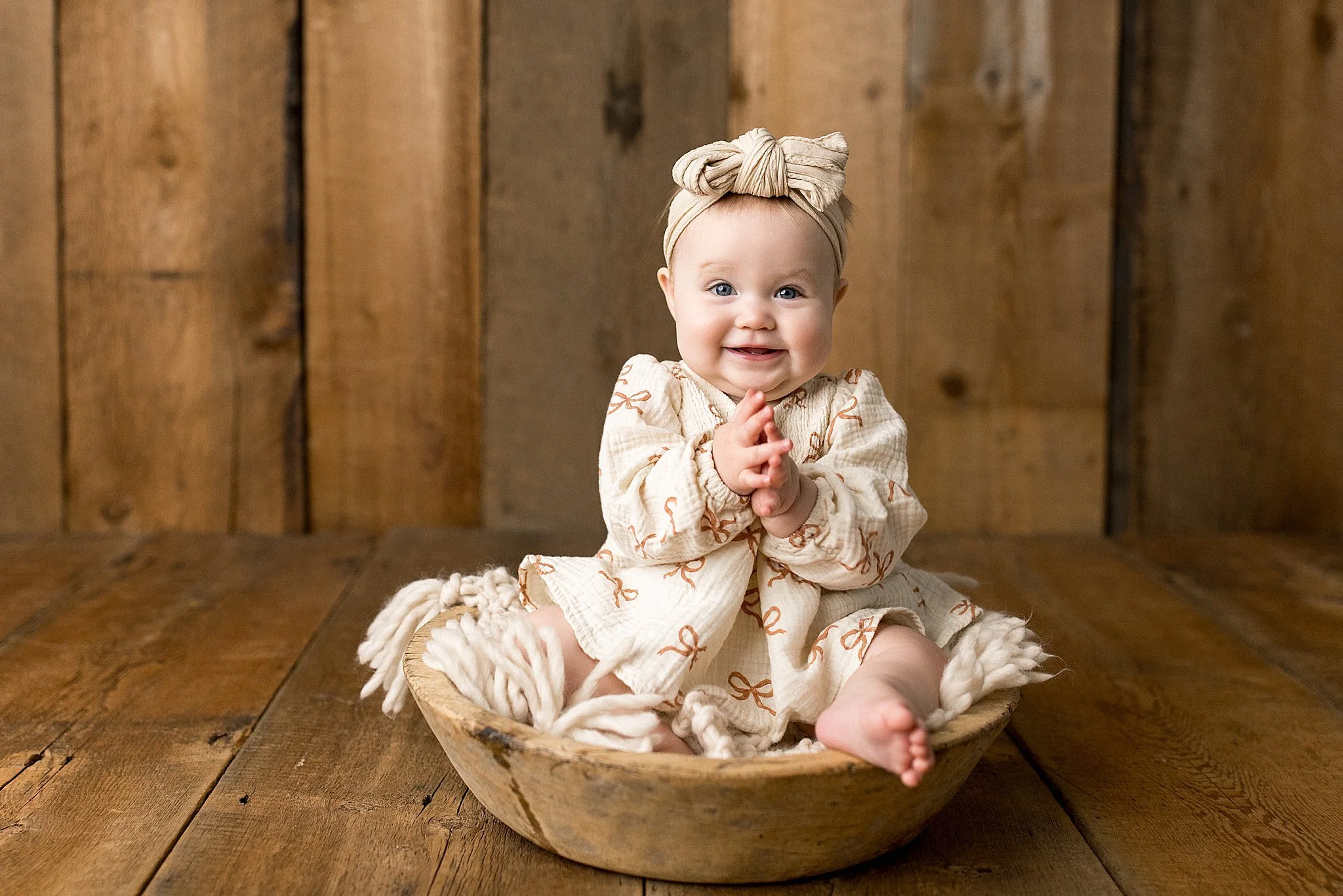 A smiling baby girl sitting in a wooden bowl on a wooden floor with a wooden background, wearing a beige dress with bow patterns and a headwrap with baby photographers near me.