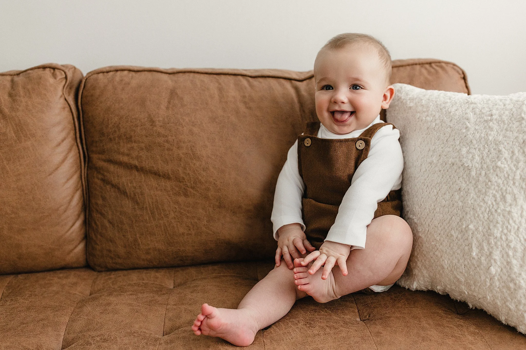 A cheerful baby sitting on a brown couch with a white fluffy pillow, wearing a white long-sleeve shirt and brown overalls with best family photographers near me.
