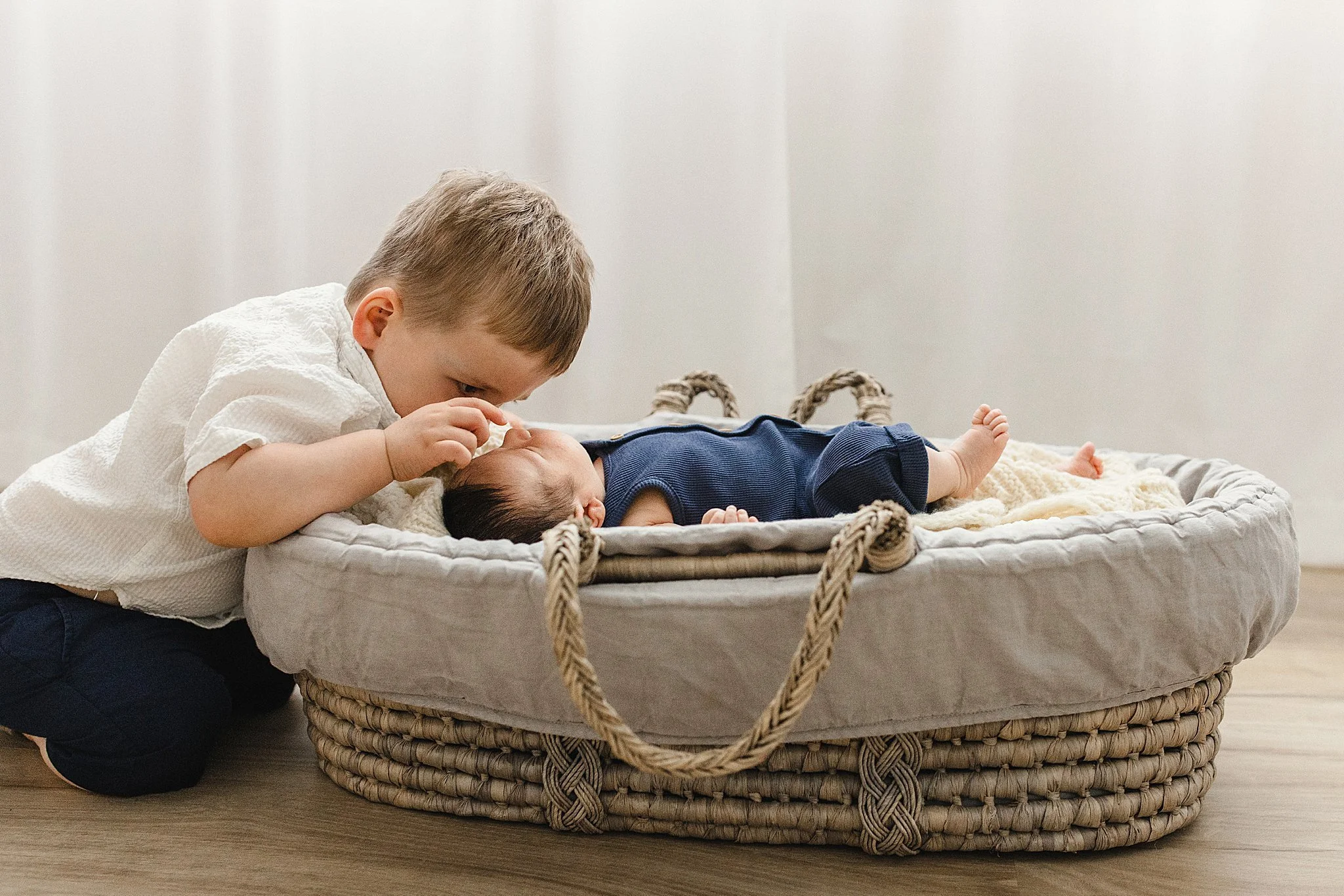 A young boy leaning over and kissing a newborn baby lying on a cushioned wicker baby bed with a lifestyle newborn photographer near me.