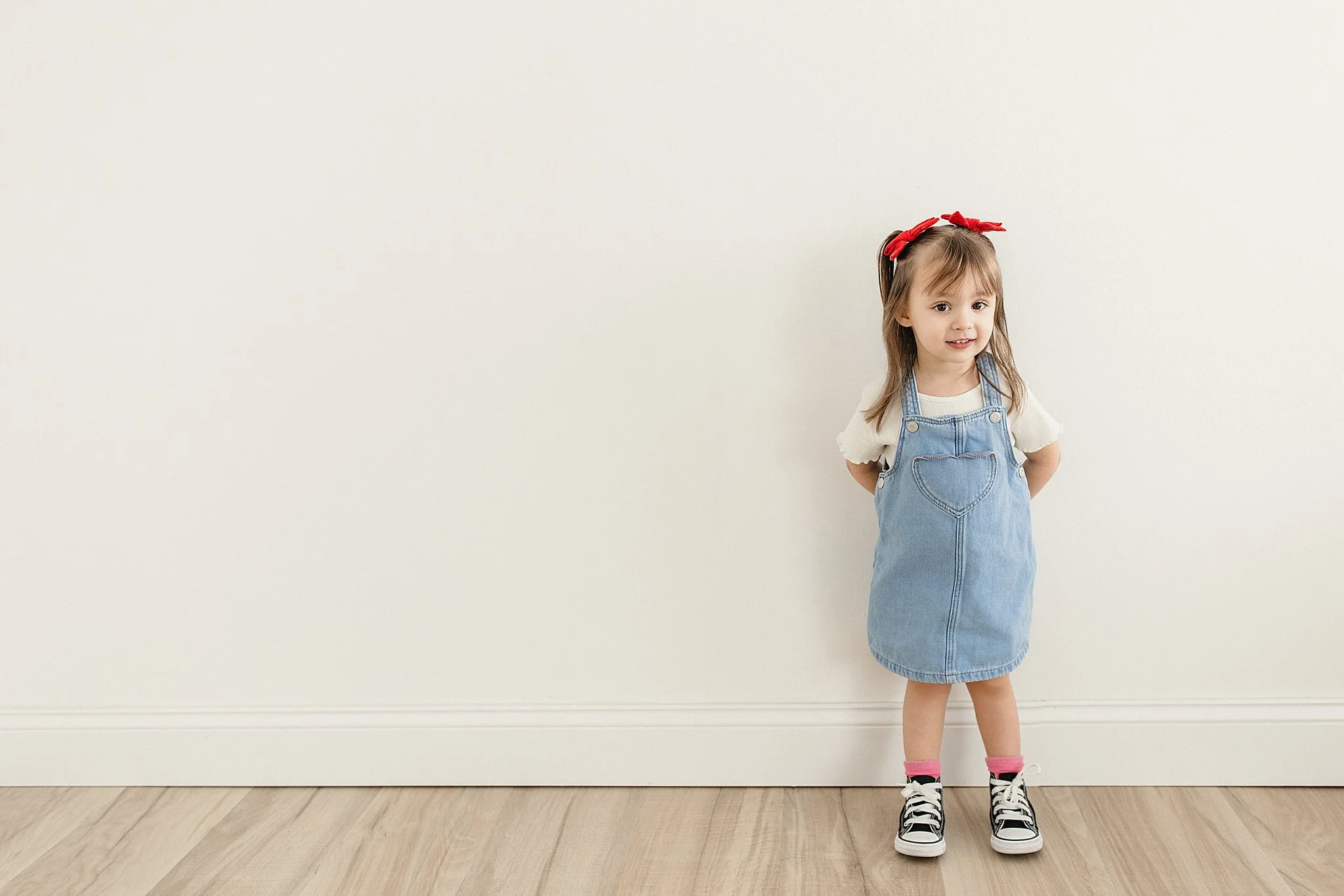 A young girl with long brown hair, wearing a red headband, a white shirt, a denim pinafore dress, pink socks, and black sneakers, standing against a plain white wall with plastic floorboards for a kids model photoshoot in Naperville, IL.