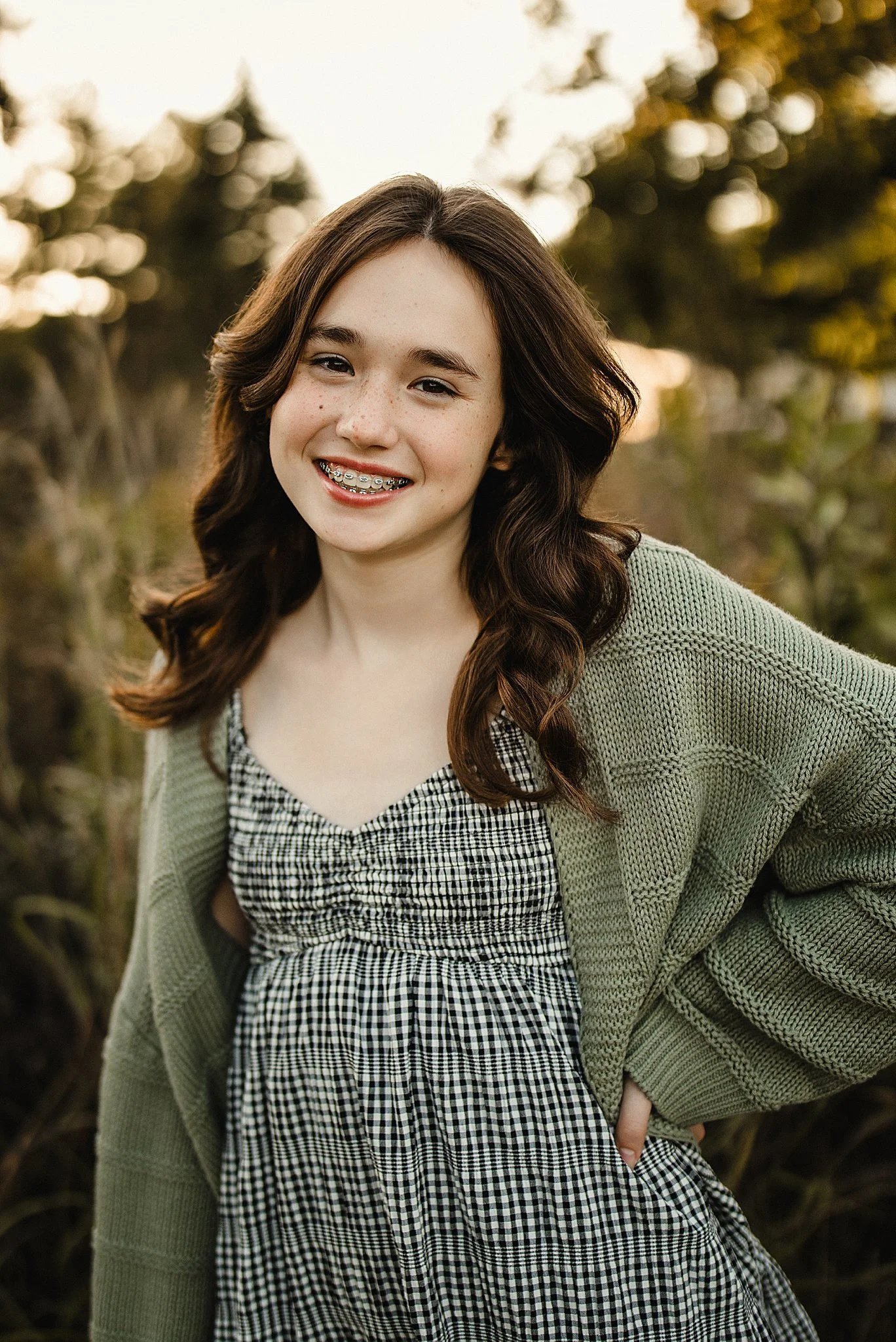A teenage girl with braces and long brown hair smiling outdoors during sunset, wearing a green cardigan over a black and white checkered dress during a Downers Grove family photo session.