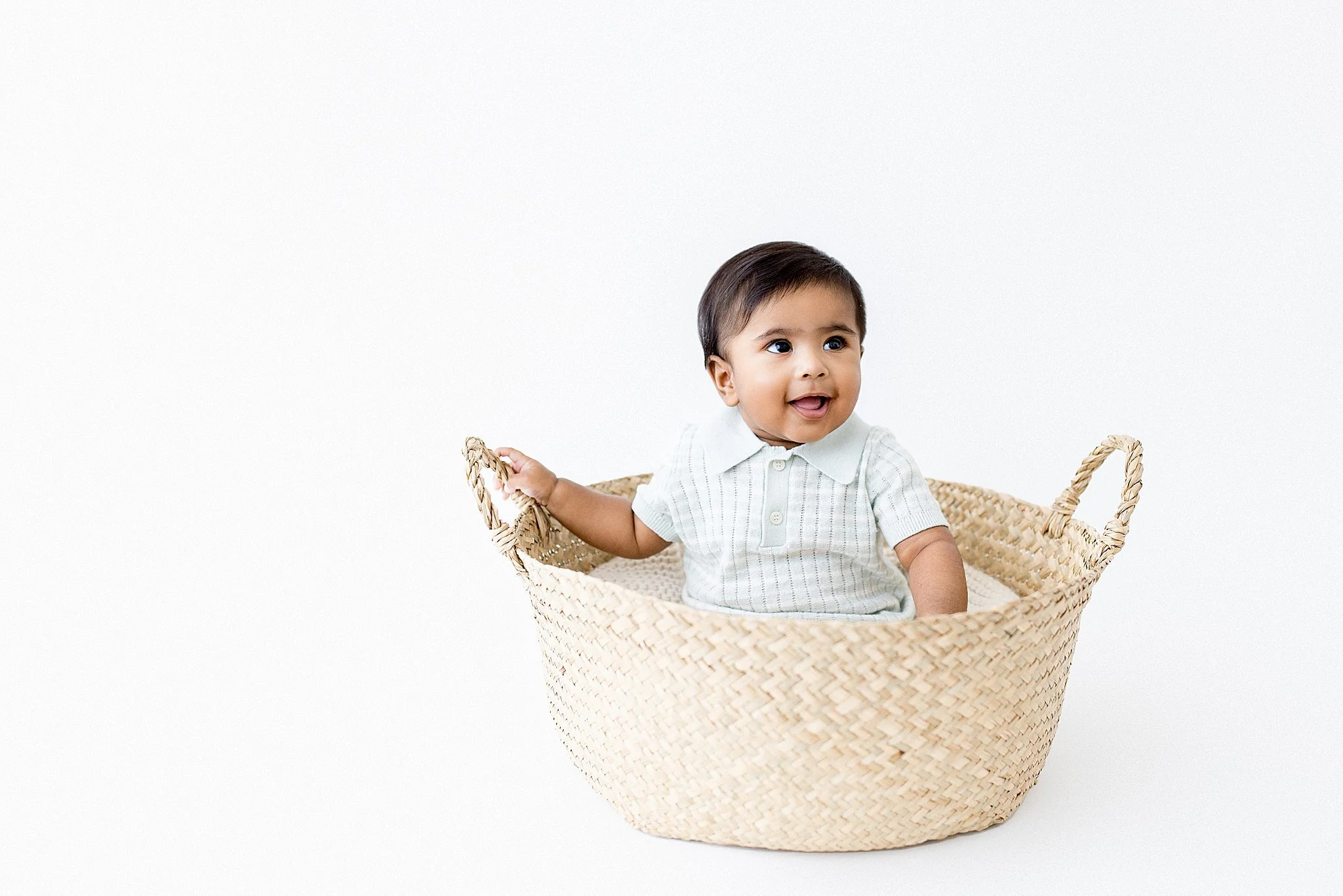 A smiling baby boy sitting in a woven basket against a plain white background for baby photographers in Naperville.