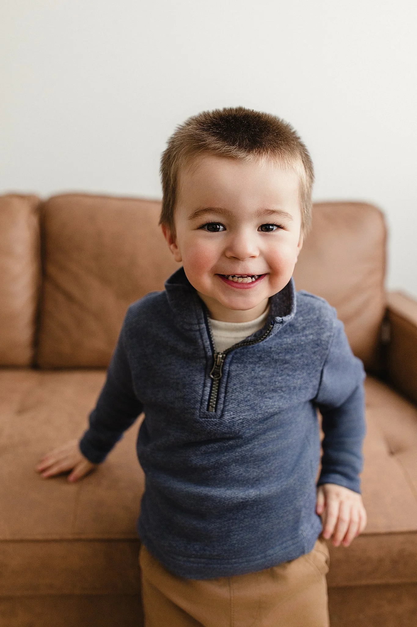 A young boy with short brown hair smiling and looking at the camera, sitting on a brown couch in a room with a plain white wall during lifestyle photos with Ally and B Photography.
