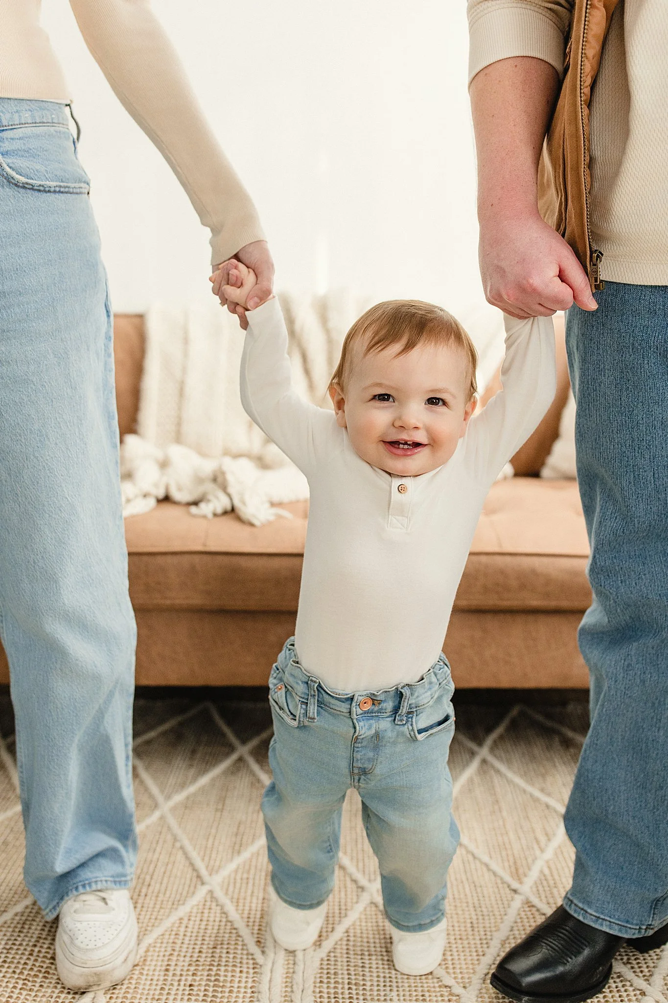 A smiling toddler holding hands with two adults, standing on a patterned rug in a living room with the best family photographer in Naperville, IL.