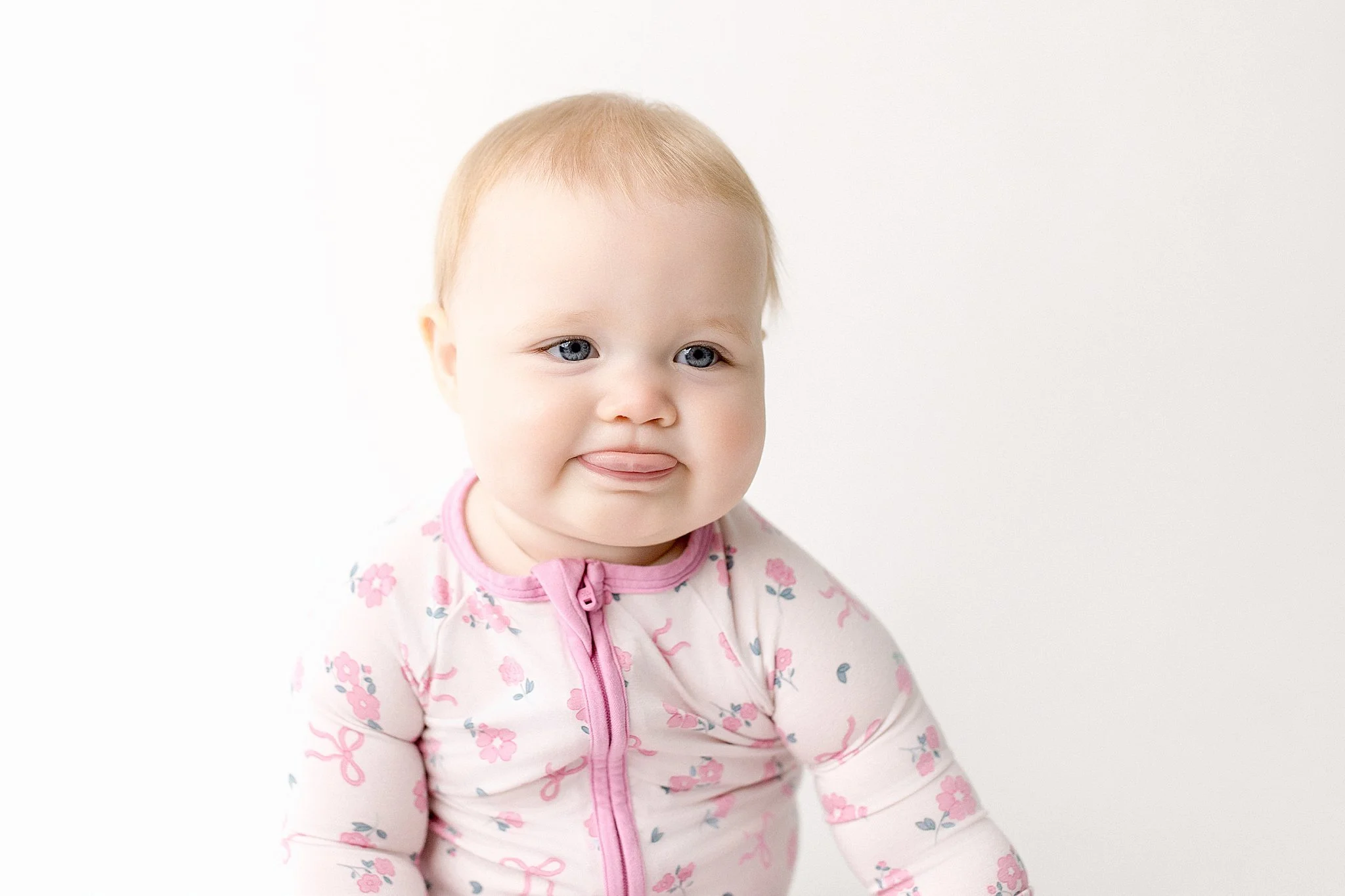 A baby girl with blonde hair and blue eyes wearing a pink and white floral jacket, sticking out her tongue, against a plain white background during a baby photoshoot in Naperville with Ally and B Photography.