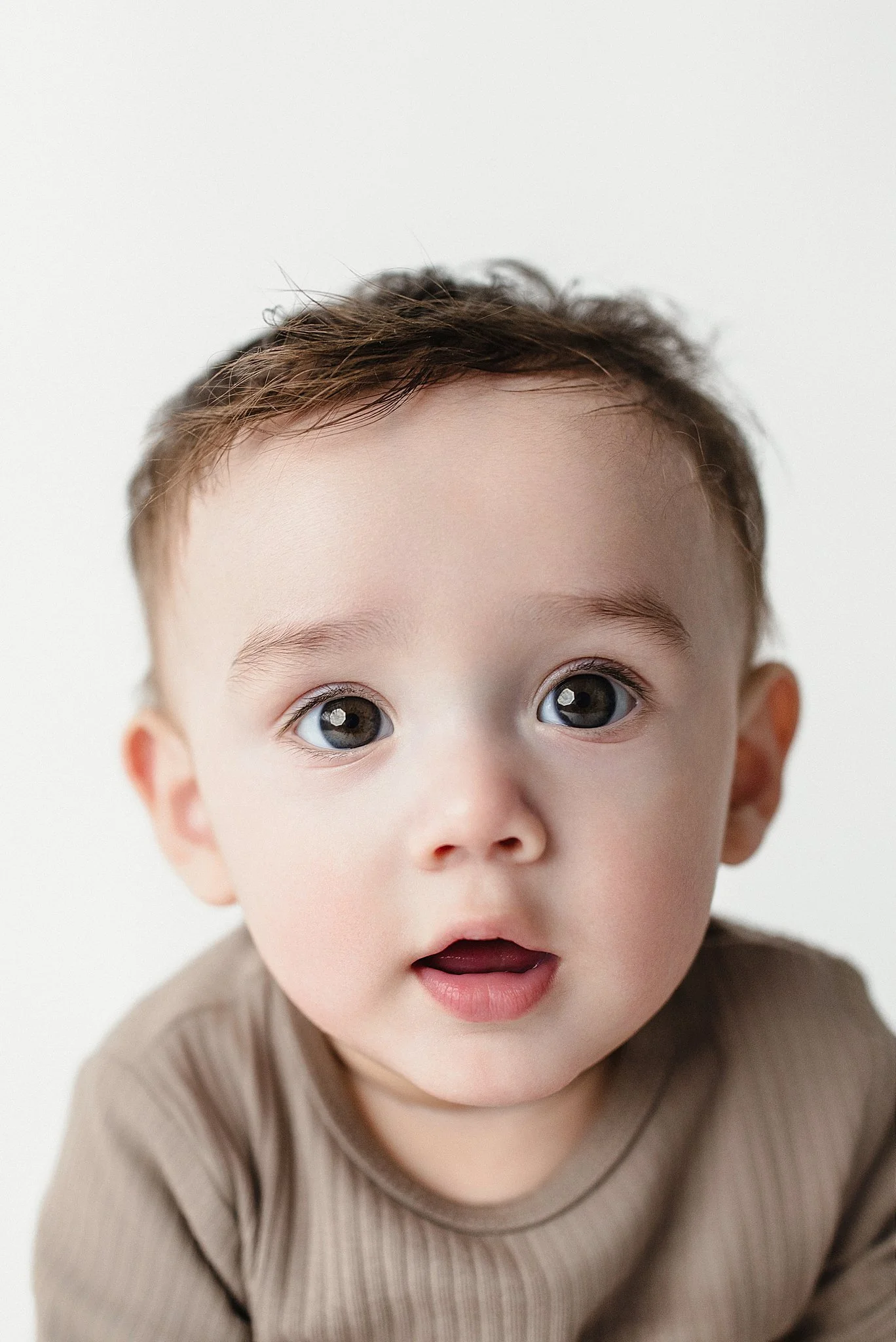 Close-up of a young child with big eyes and a surprised expression, wearing a beige shirt against a white background.