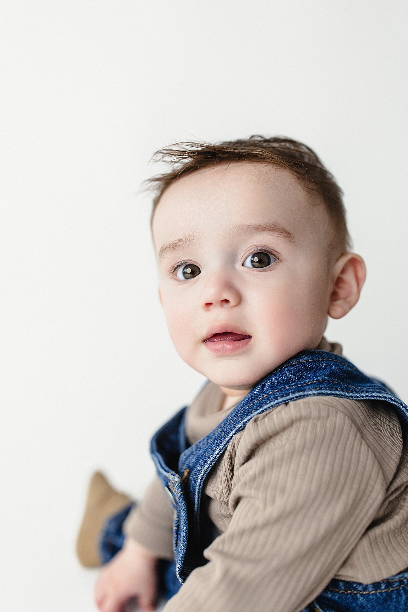 Close-up of a young boy with wide eyes, wearing a beige long-sleeve shirt and denim overalls, against a plain white background.