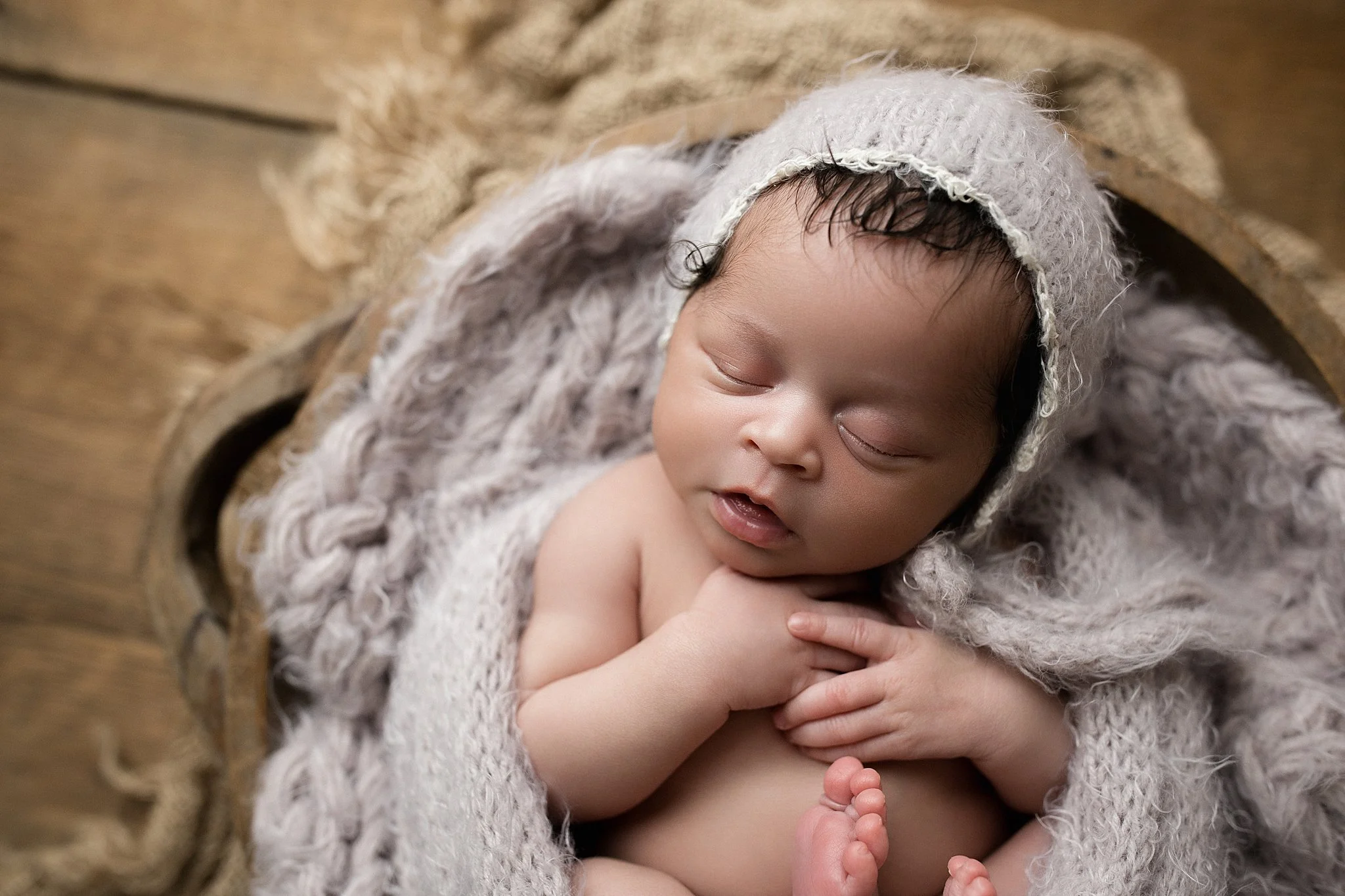 A peacefully sleeping baby wearing a knitted hat and blanket, lying on a wooden surface during posed newborn photography near me.